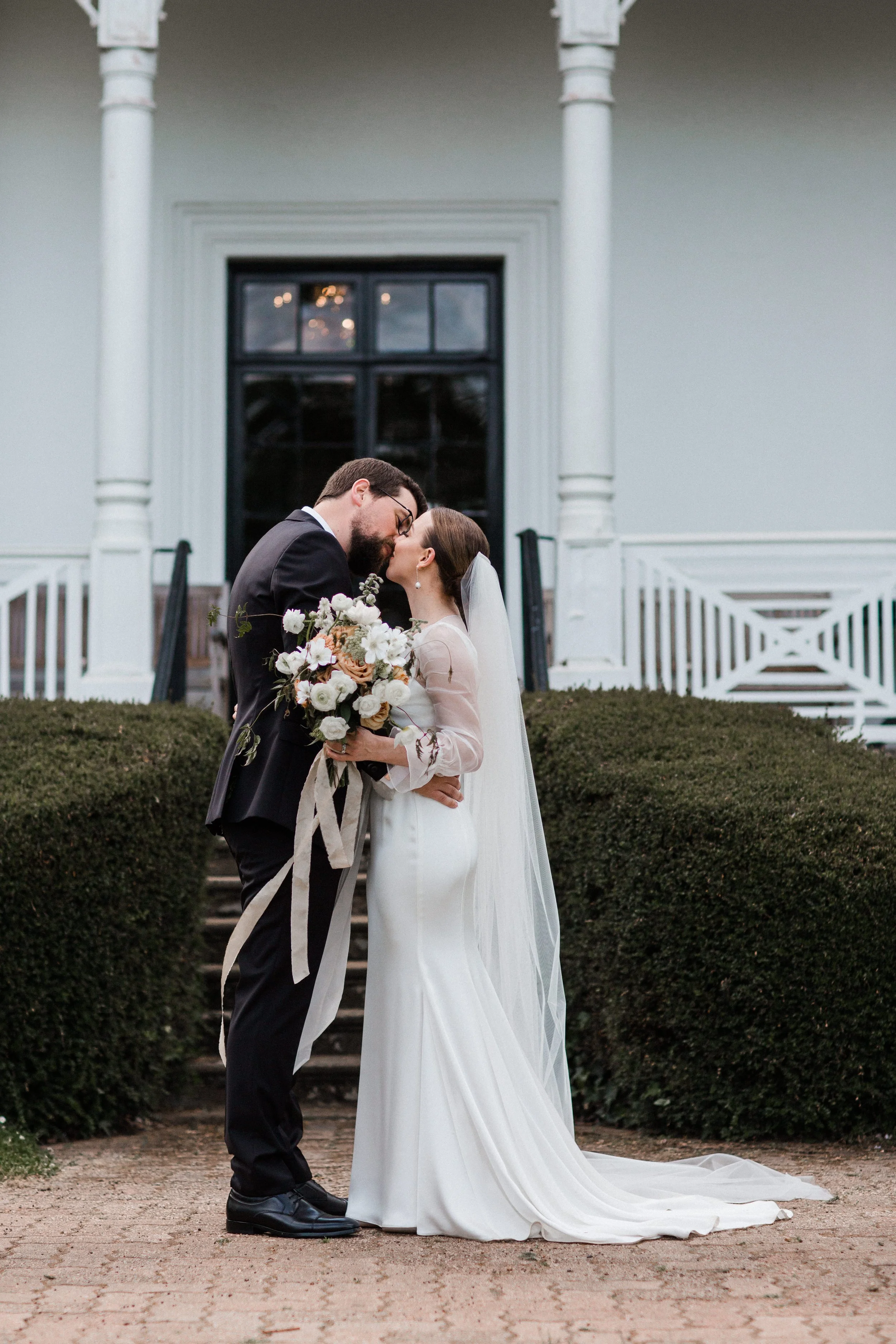 A newlywed couple sharing a kiss outdoors in front of a white house, with the bride holding a bouquet of white and blush-colored flowers.