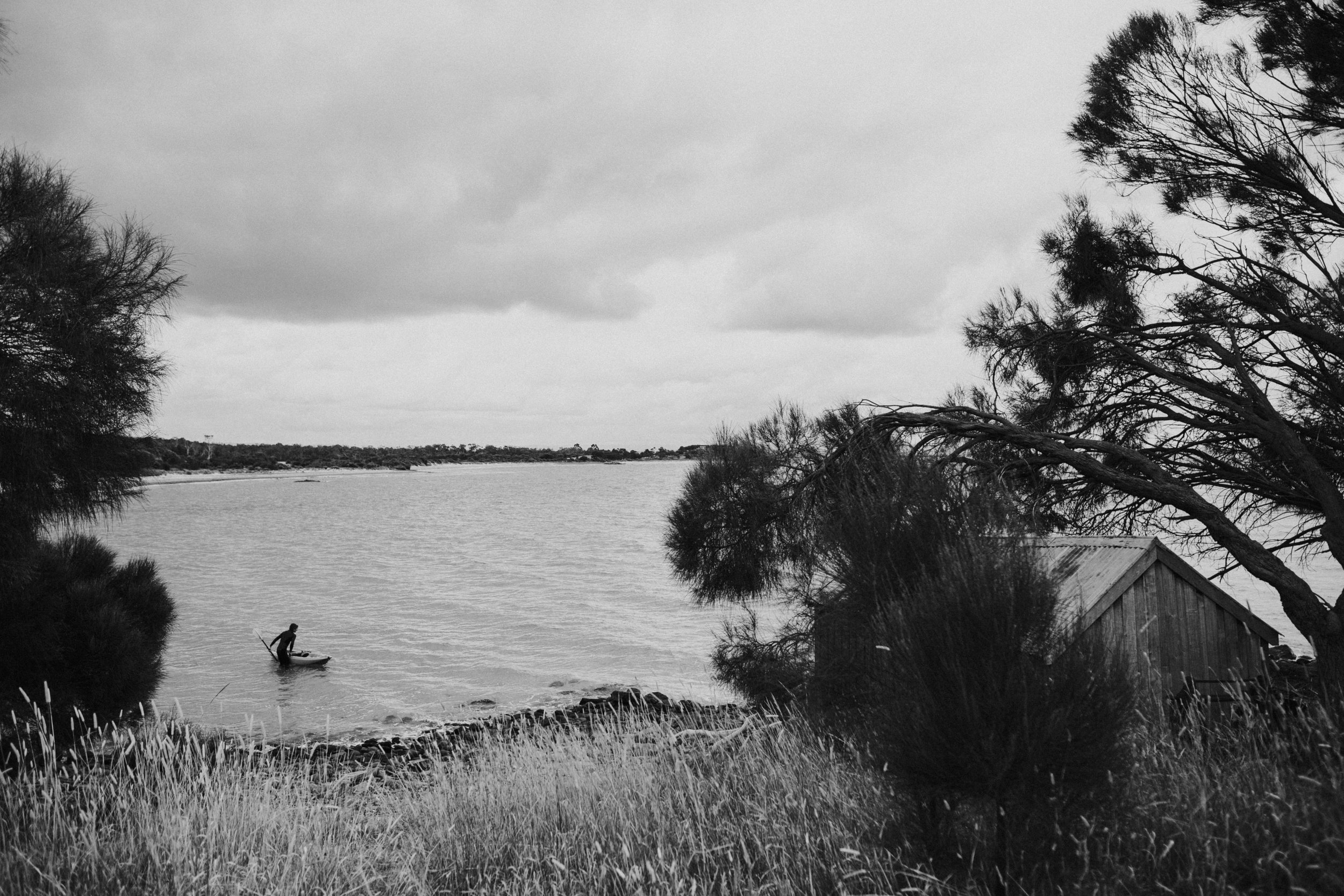 A black and white photo of a lakeside scene with a person standing on a paddleboard on the water. There are trees on the right and left sides, a small wooden structure near the water, and a cloudy sky overhead.