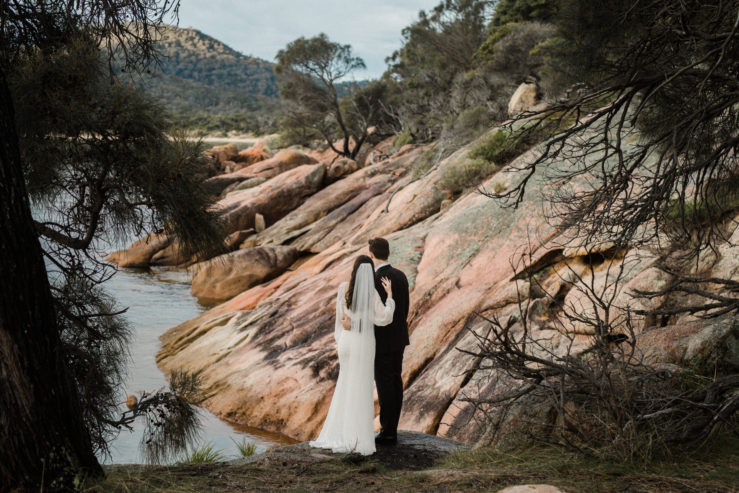 A bride and groom dressed in wedding attire standing by a lakeside, surrounded by rocks and trees, with a scenic mountainous landscape in the background.