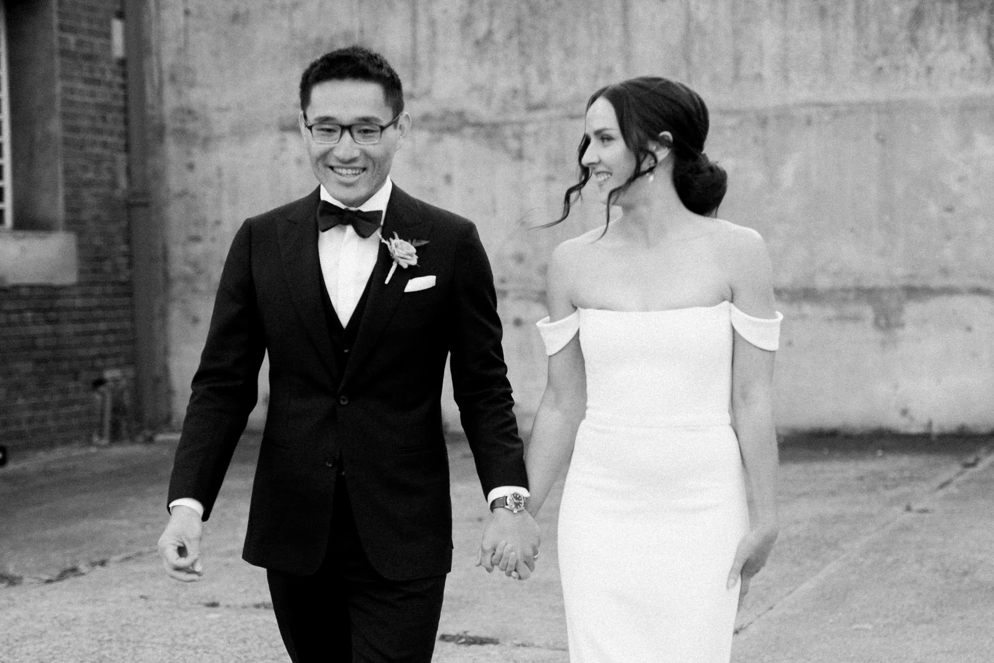 A smiling man and woman in wedding attire holding hands and walking outdoors against a concrete wall, black and white photo.