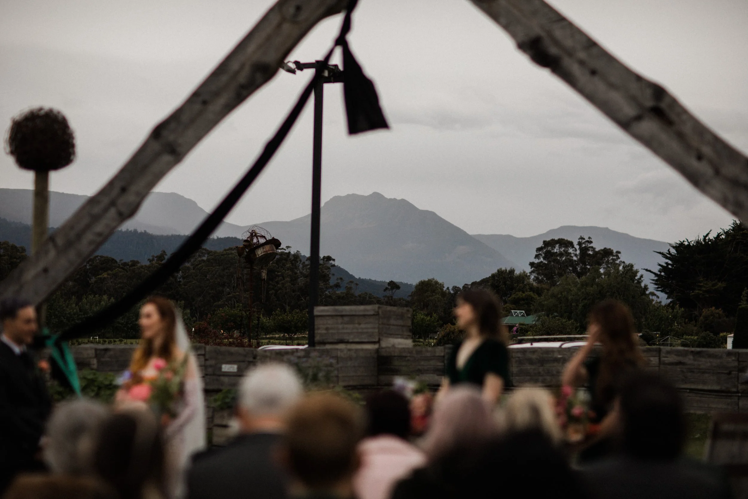 A wedding ceremony outdoors with mountains in the background, people gathered, a bride in a white wedding dress, a groom in a dark suit, and attendees, with a wooden structure and cloudy sky overhead.