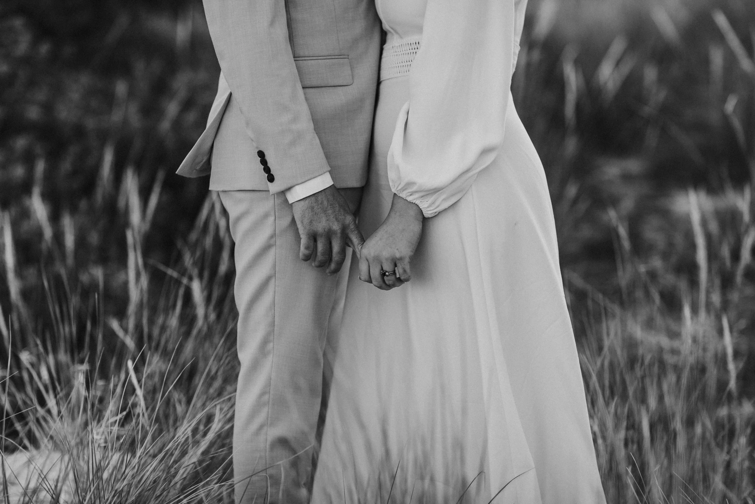 A close-up black and white photo of a couple holding hands, with the woman in a long dress and the man in a suit, standing in a natural outdoor setting.