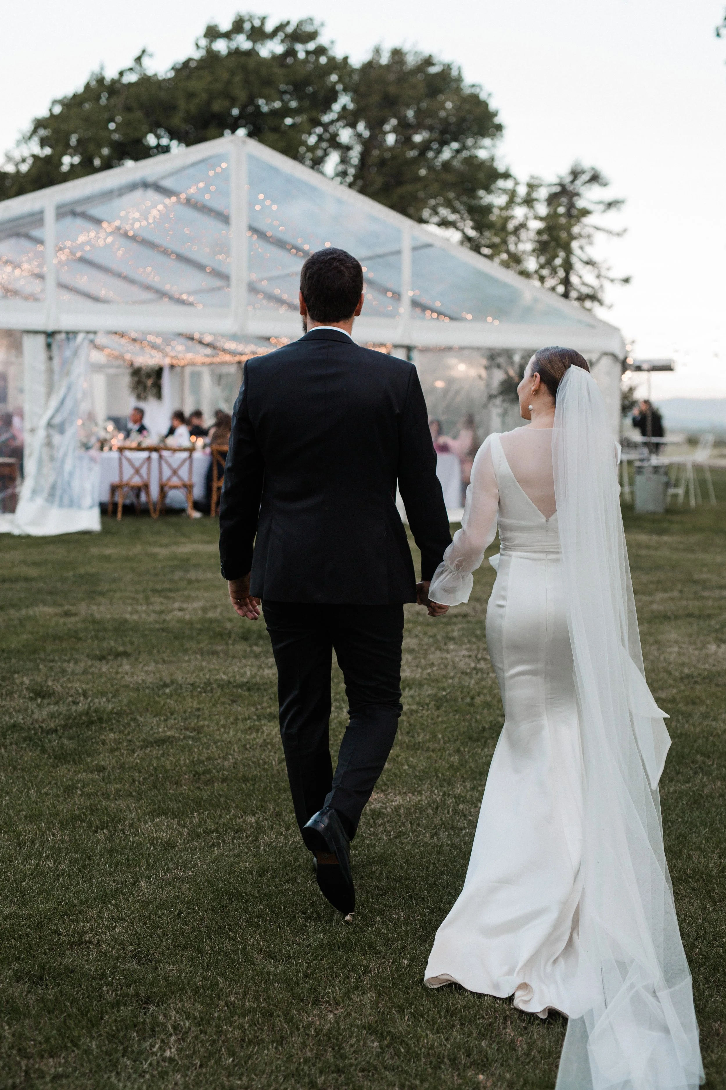 A bride and groom walking hand in hand outside towards a decorated reception tent, with guests seated inside.