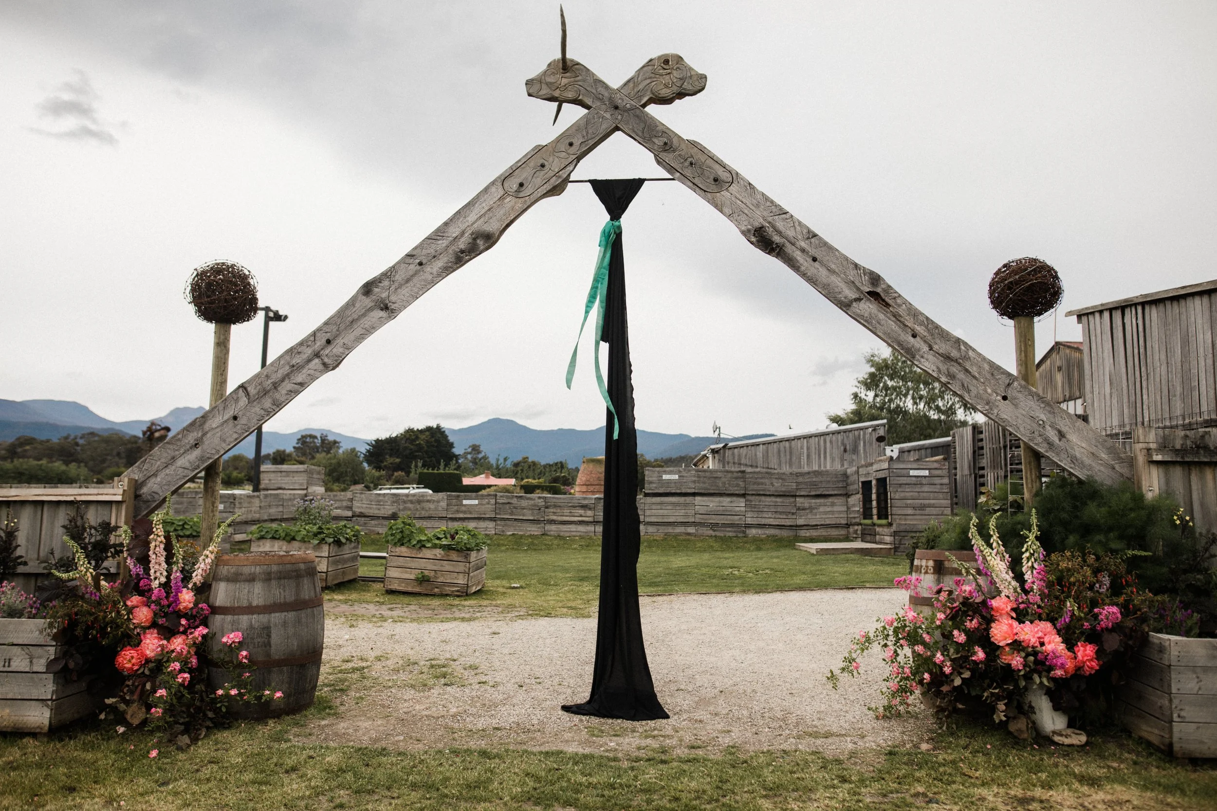 A rustic outdoor wedding arch made of weathered wooden beams with decorative horse heads and green ribbons, surrounded by flower arrangements and wooden barrels, with mountains in the background under a cloudy sky.