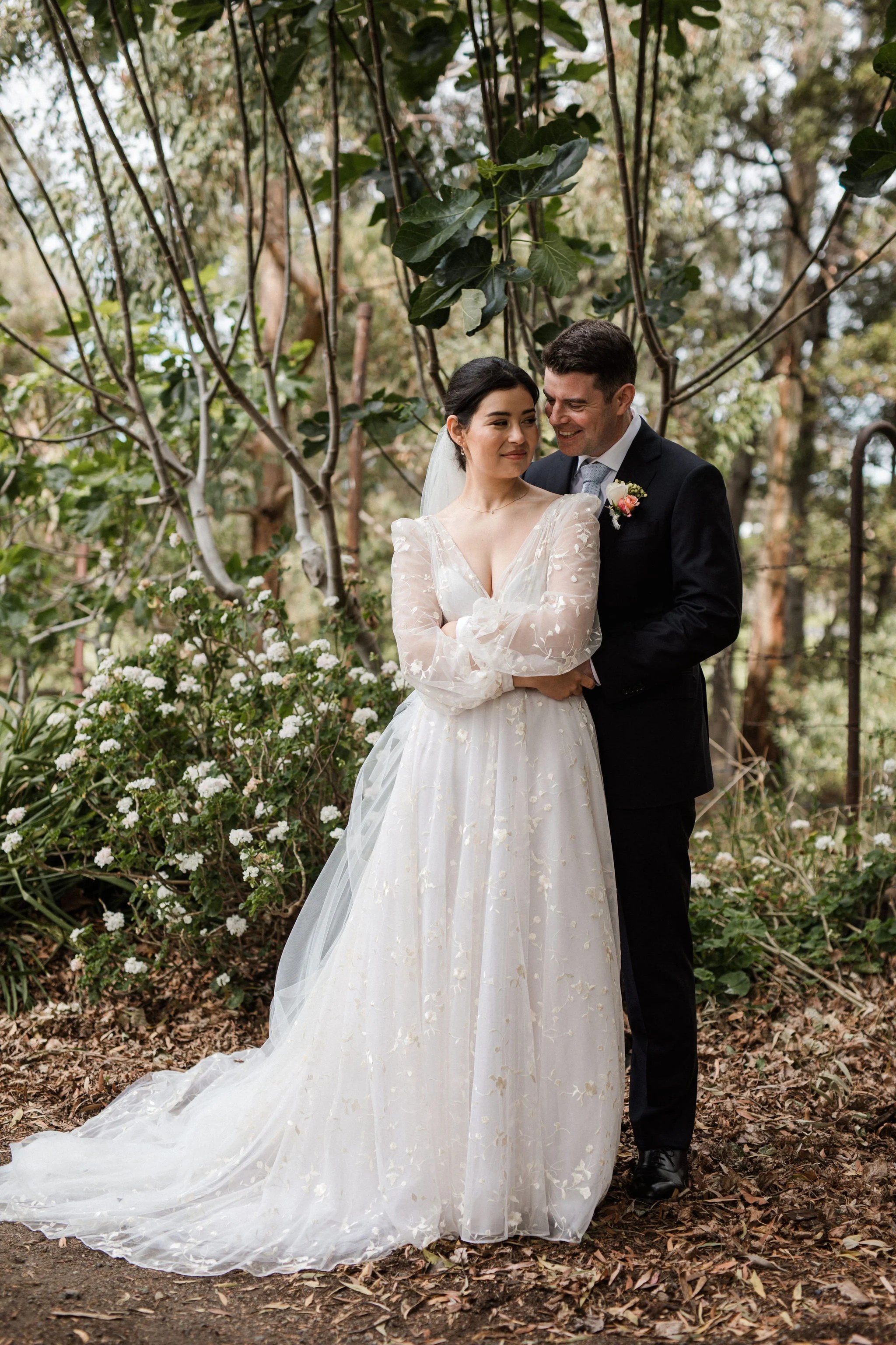 A bride and groom standing close together outdoors among trees and plants, with the bride wearing a long white wedding dress with sheer sleeves and the groom in a black suit with a boutonniere, both smiling softly.