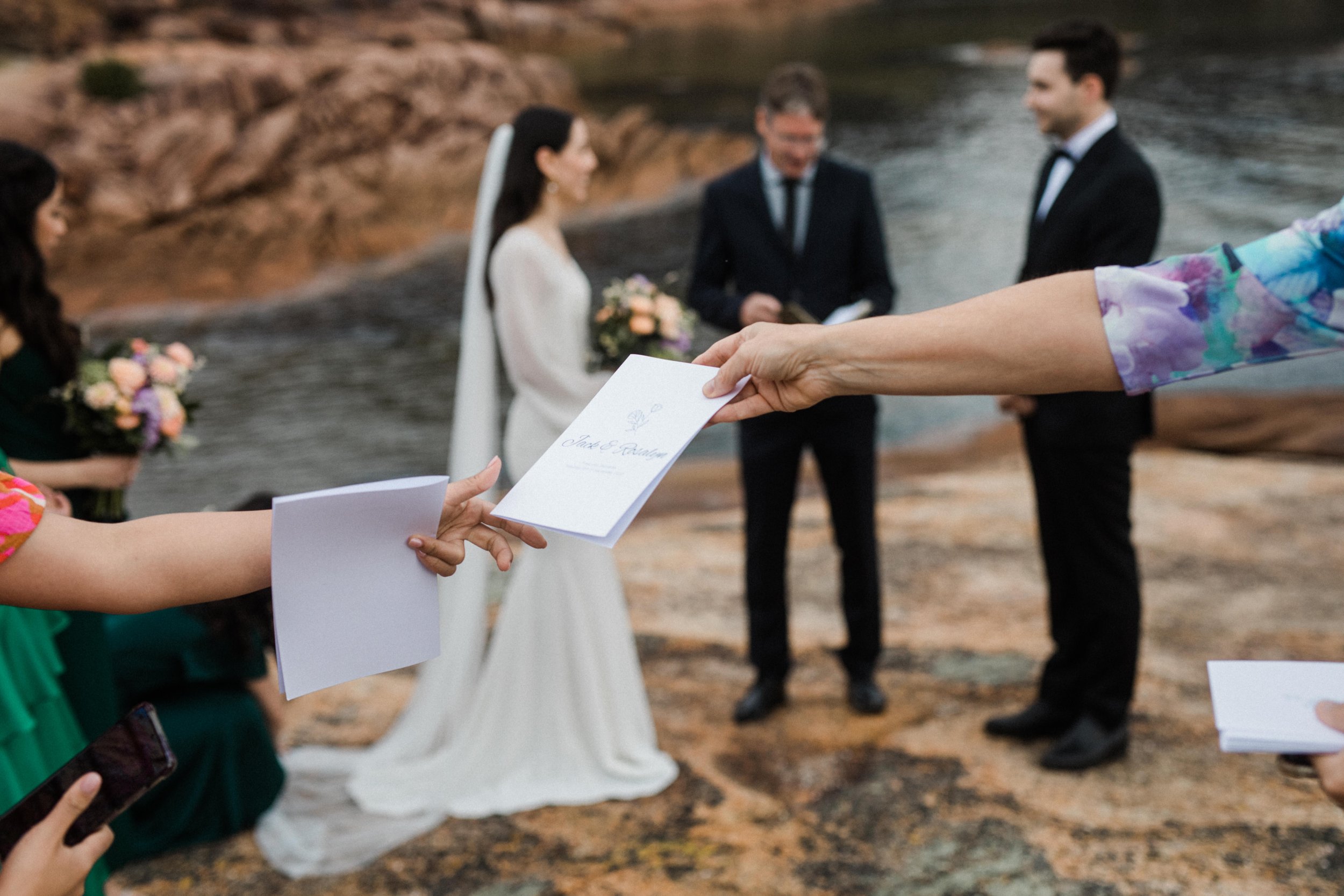 Close-up of a wedding ceremony with a person handing a wedding vow booklet to another person, with the bride and groom in the background near water and rocks.