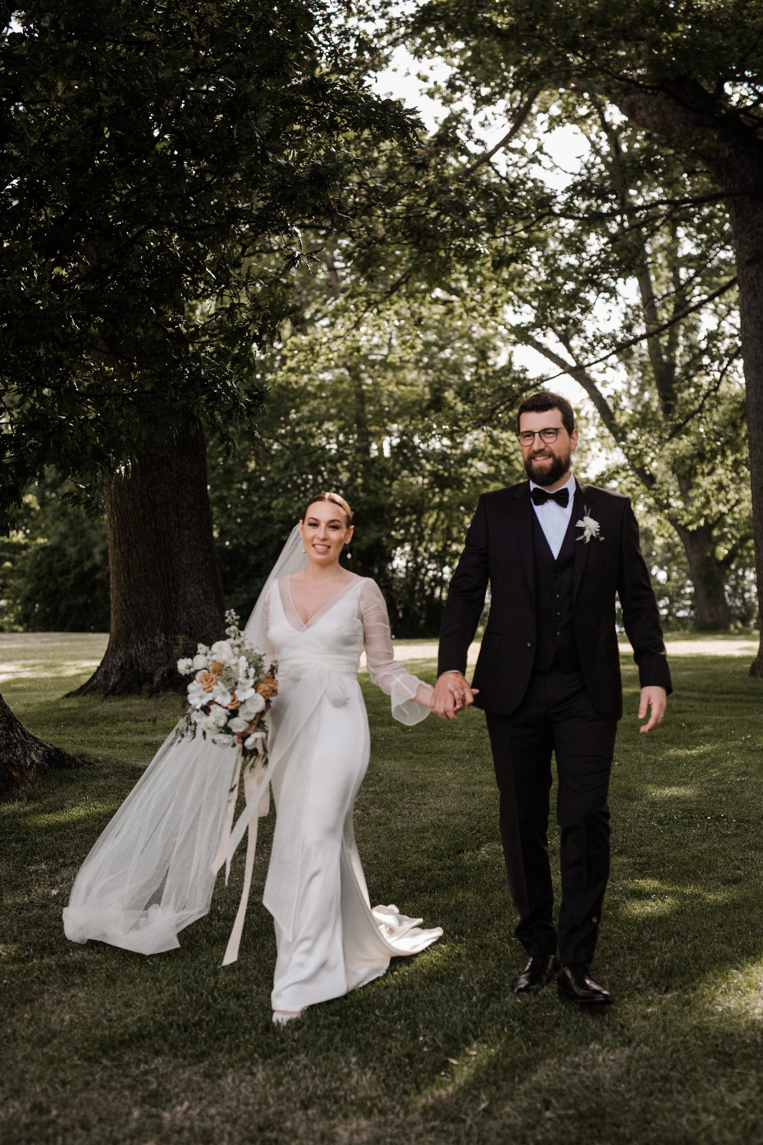 A bride and groom holding hands and walking outdoors on a grassy area with large trees in the background on their wedding day.