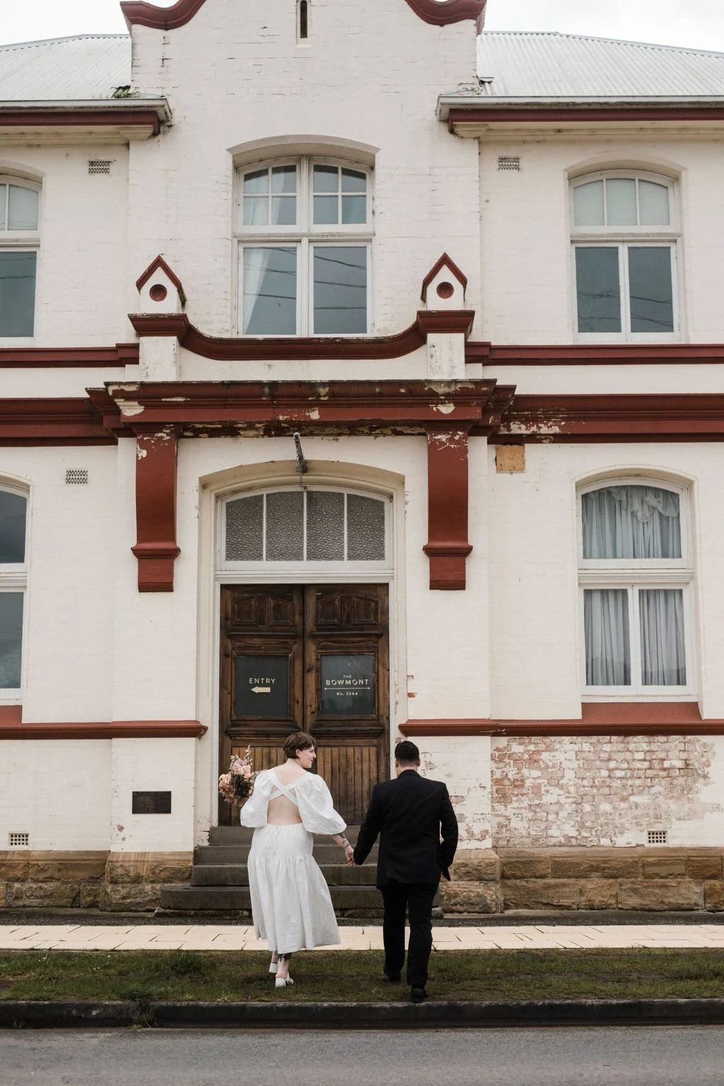 A couple dressed in wedding attire walking hand in hand towards an old building with a wooden door, white brick walls, and large windows.