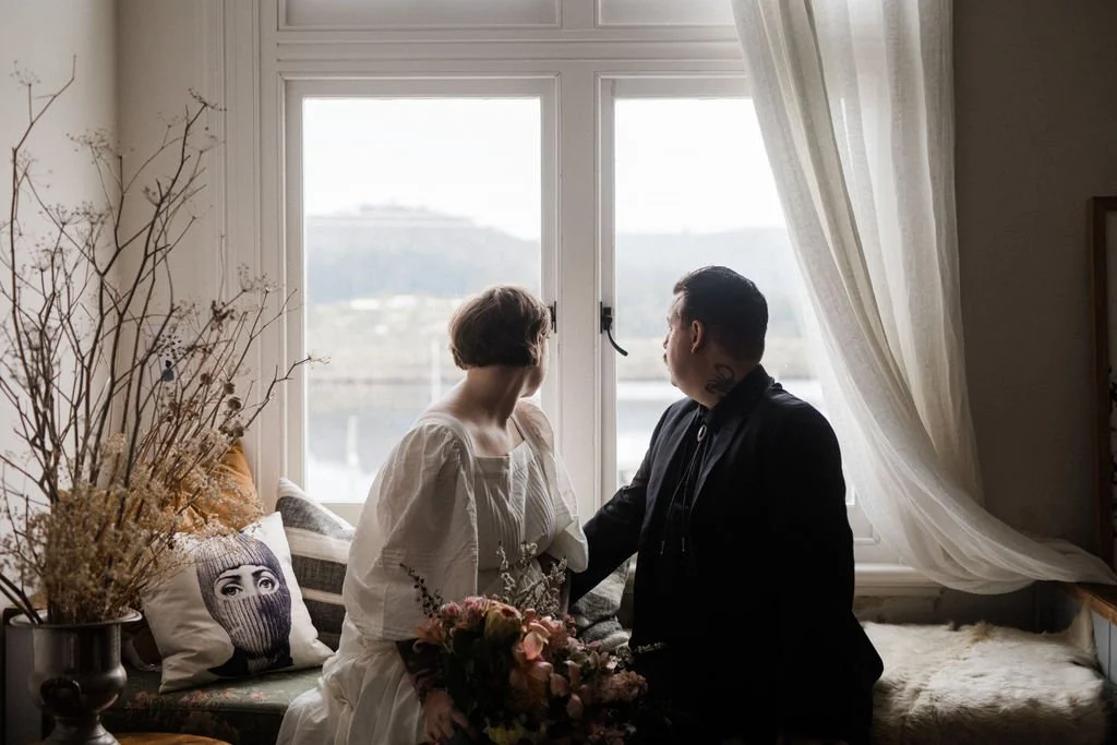 A couple is sitting on a window seat, holding hands and looking out the window, with a bouquet of flowers in the woman's lap. The room is decorated with dried flowers and pillows, and sheer curtains drape by the window.