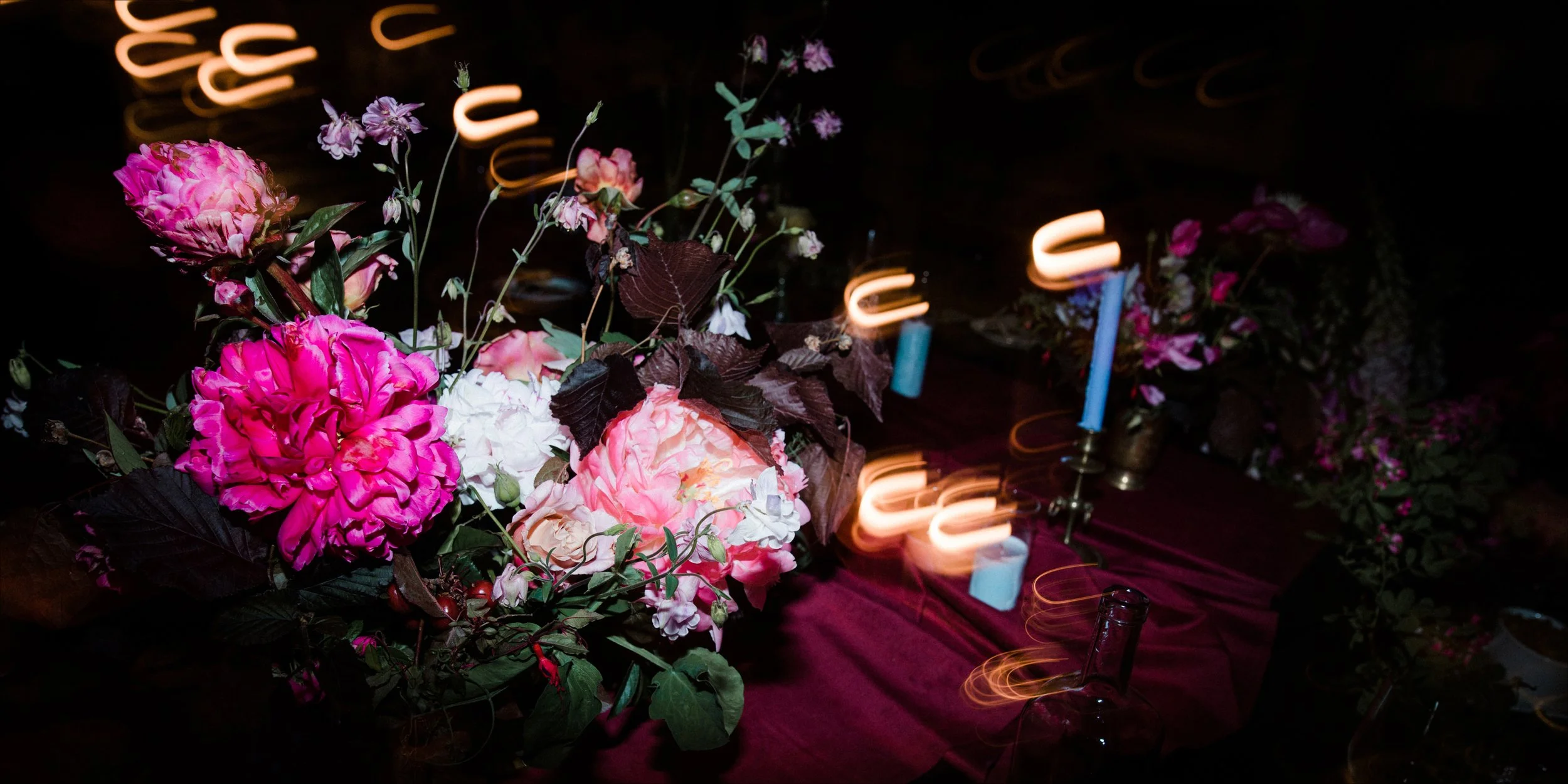 Pink and white flowers arranged on a table with a burgundy cloth, candles, and dark background