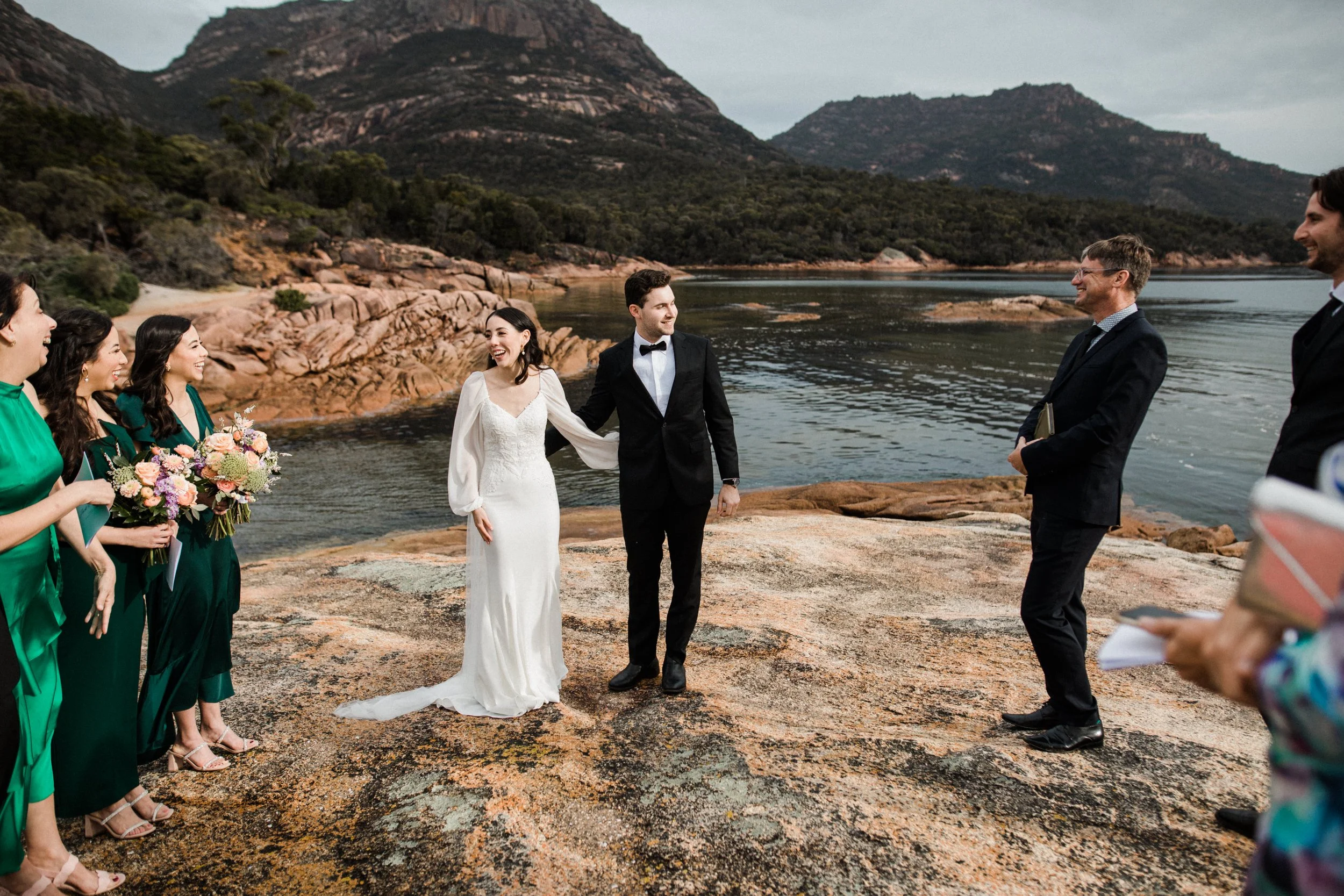 A wedding ceremony taking place outdoors on a rocky surface near a body of water with mountains in the background. The bride and groom are standing together, holding hands, surrounded by bridesmaids in green dresses holding bouquets and groomsmen in 