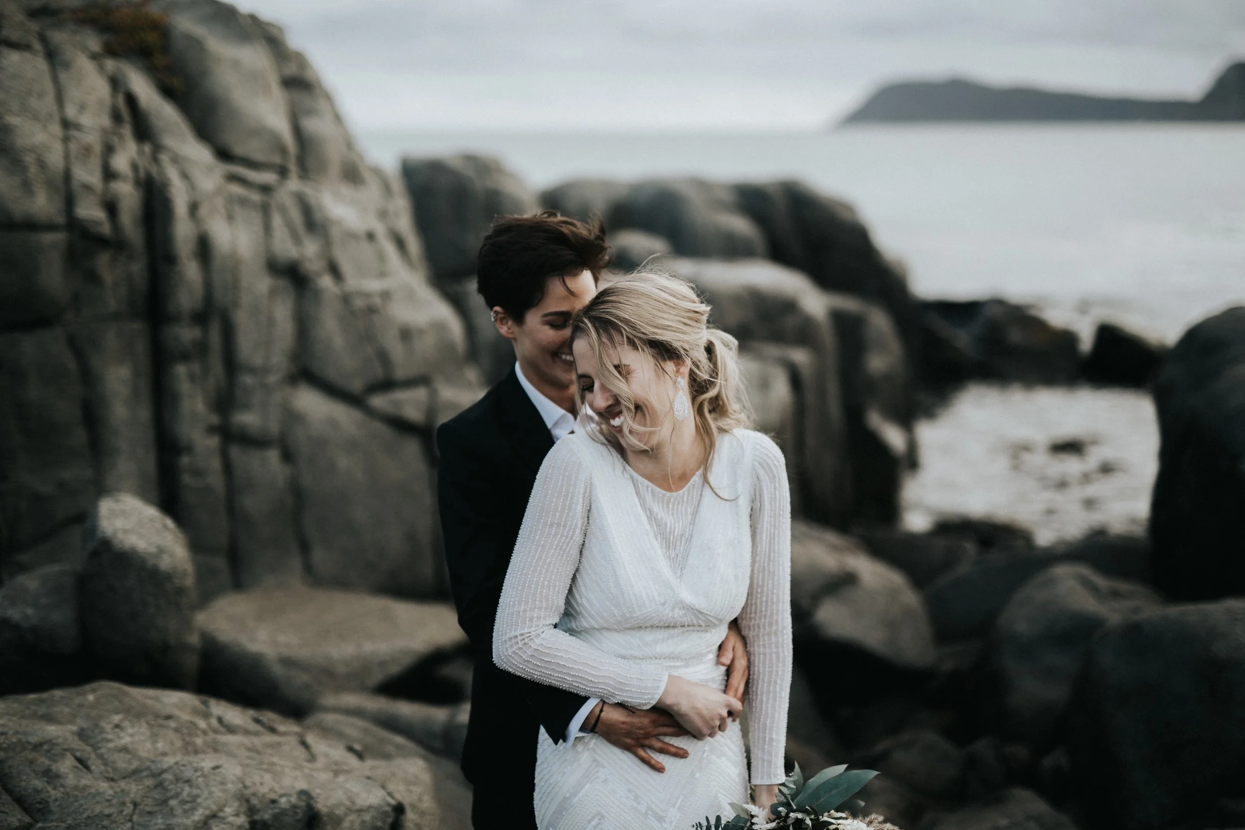 A couple in wedding attire sharing a joyful moment on a rocky beach.