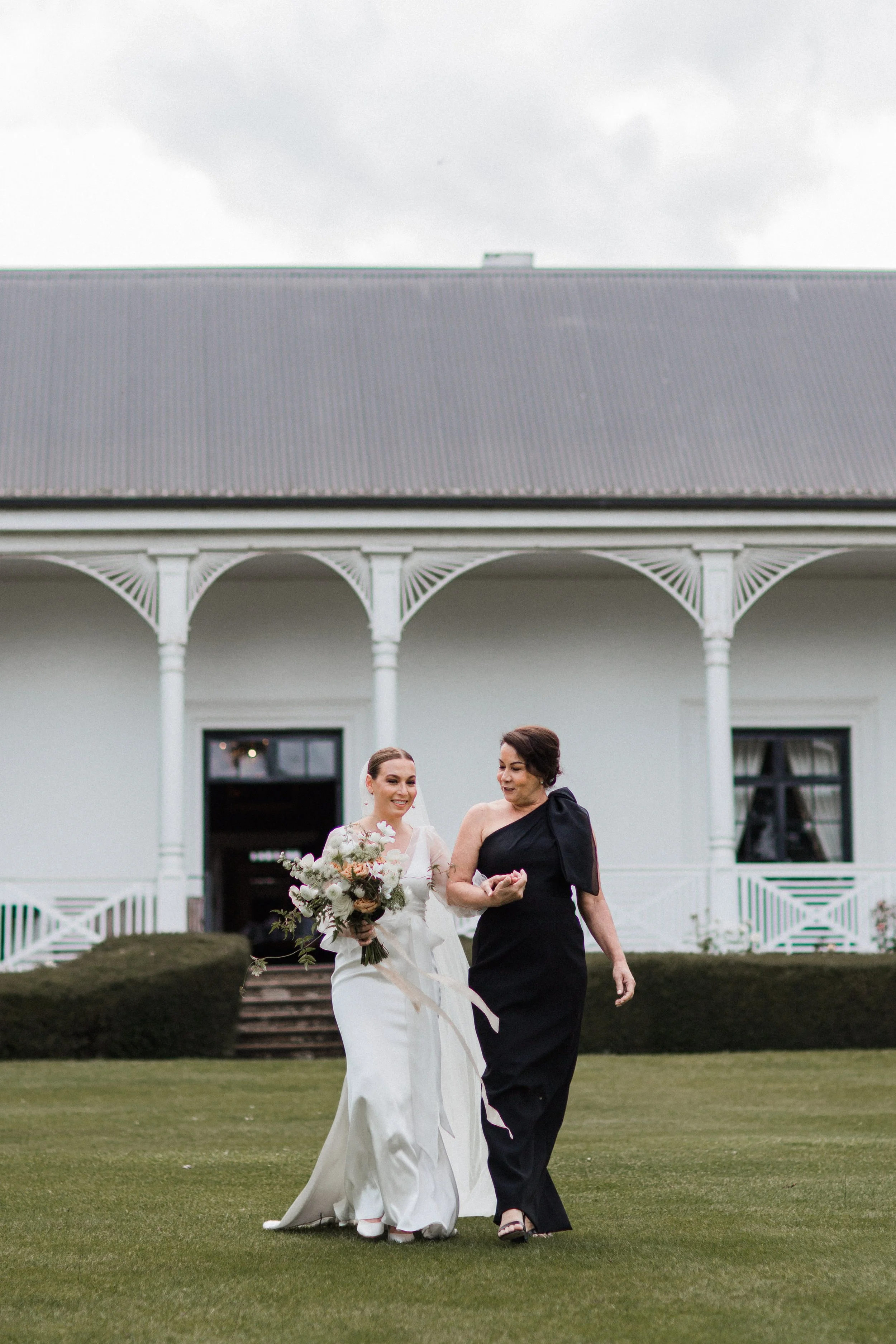 A bride in a white wedding dress walking with a woman in a black evening gown on a lawn in front of a large white house.