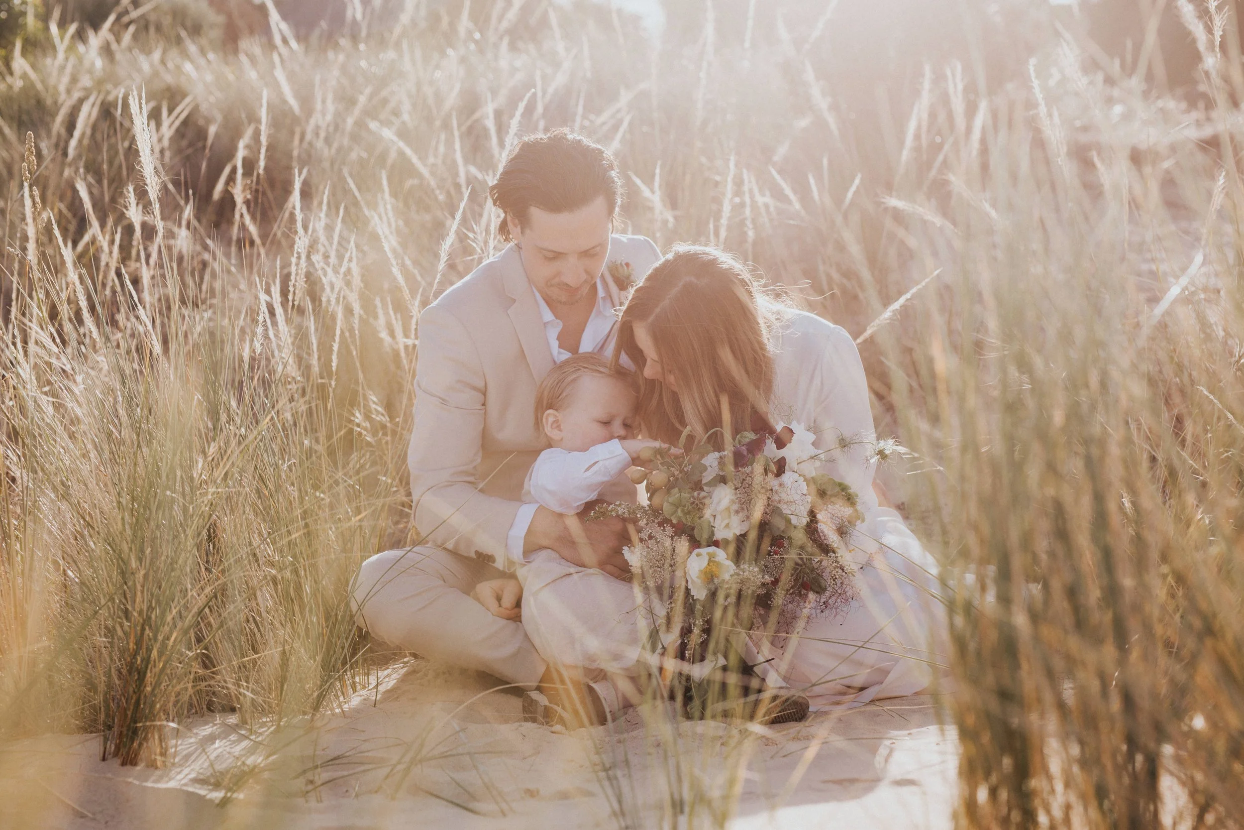 A family of three in a grassy field with tall grass, sitting on the ground, close together, with a bouquet of flowers in front of them, all leaning in towards the child.