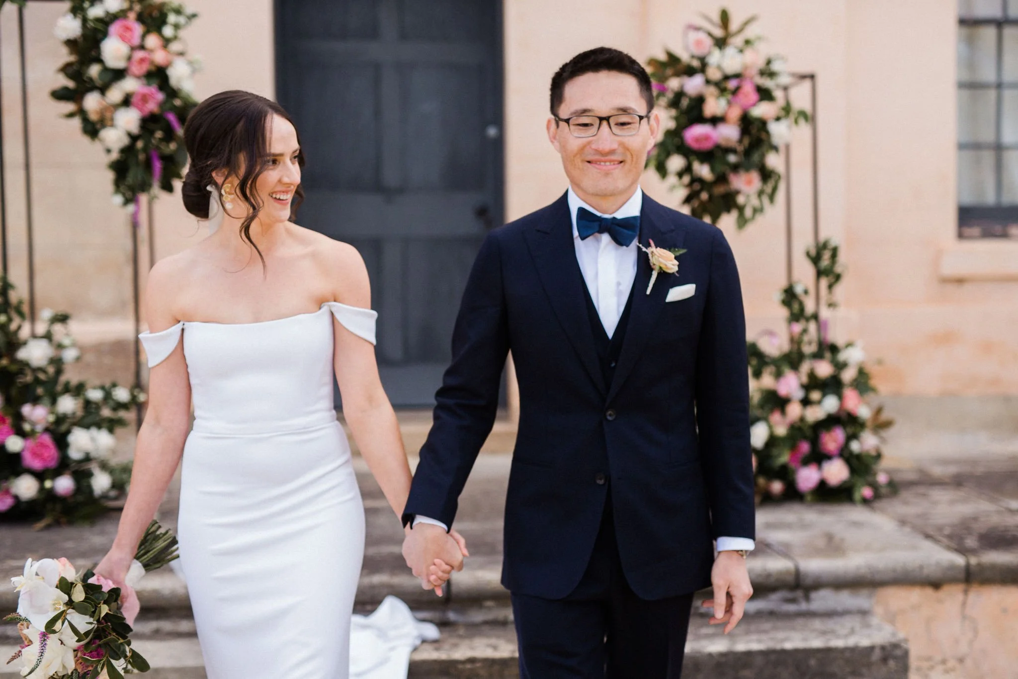A bride and groom walking hand in hand outdoors during their wedding ceremony, smiling, with floral arrangements in the background.