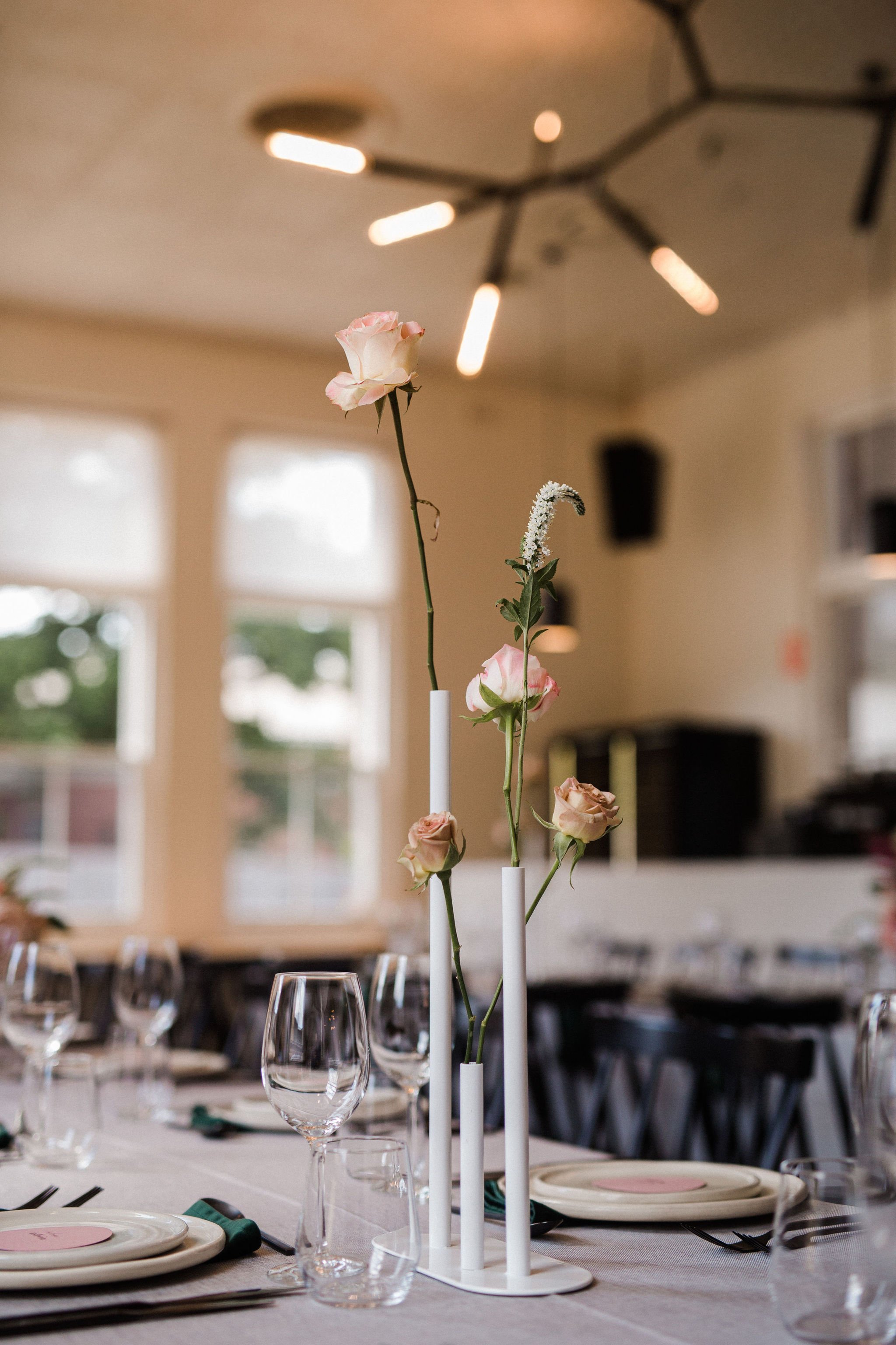 Elegant table setting with a contemporary floral centerpiece featuring pink roses and white flowers in white candleholders, set with wine glasses, plates, and utensils in a well-lit room with large windows.