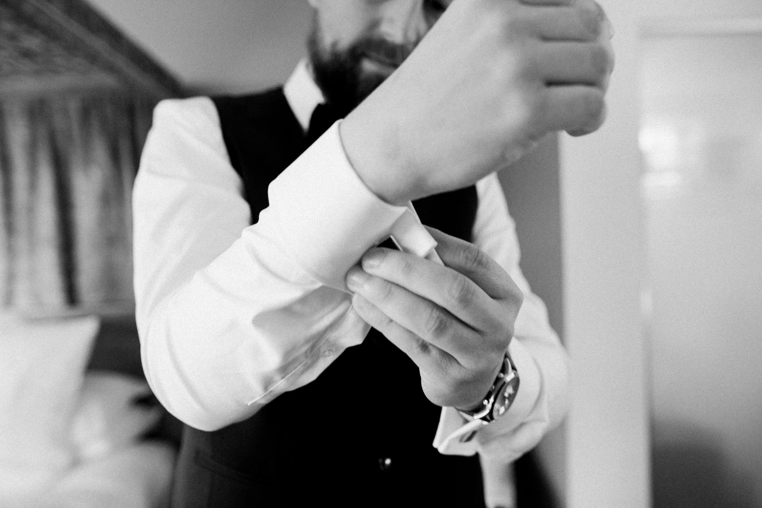 Man adjusting his sleeve in a formal setting, wearing a dress shirt, vest, and watch, with blurred background.