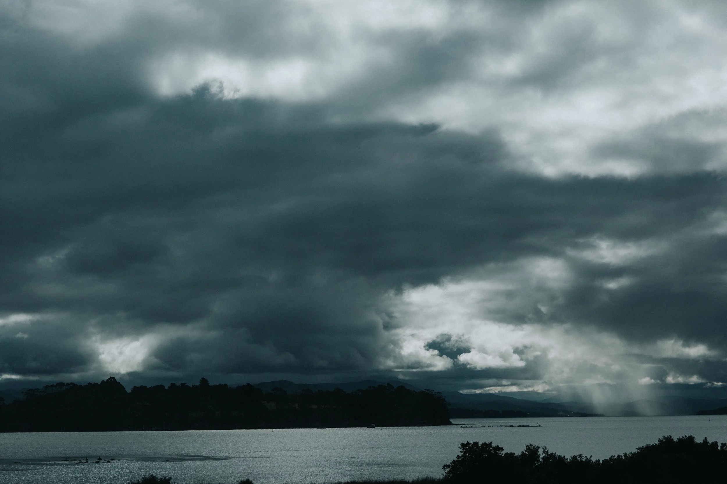 Dark storm clouds over a body of water with a forested shoreline and distant hills.
