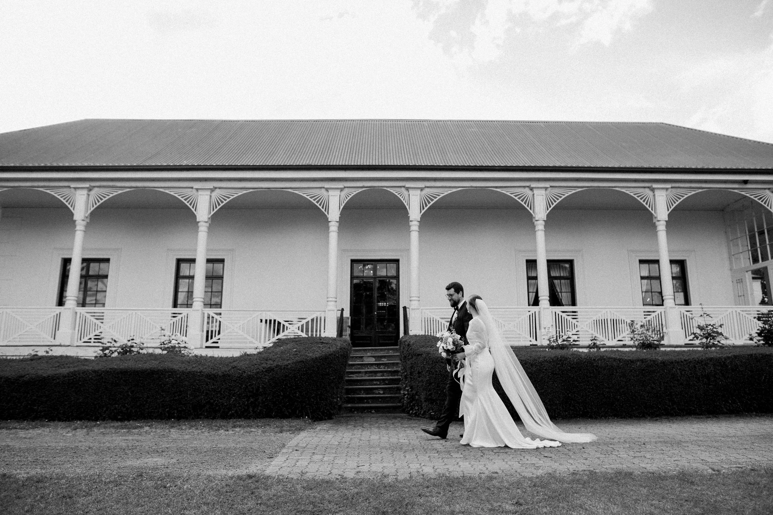 A bride and groom walking in front of a large, white, historic building with a porch supported by decorative columns, black windows, and steps leading up to the entrance, on a cobblestone path.