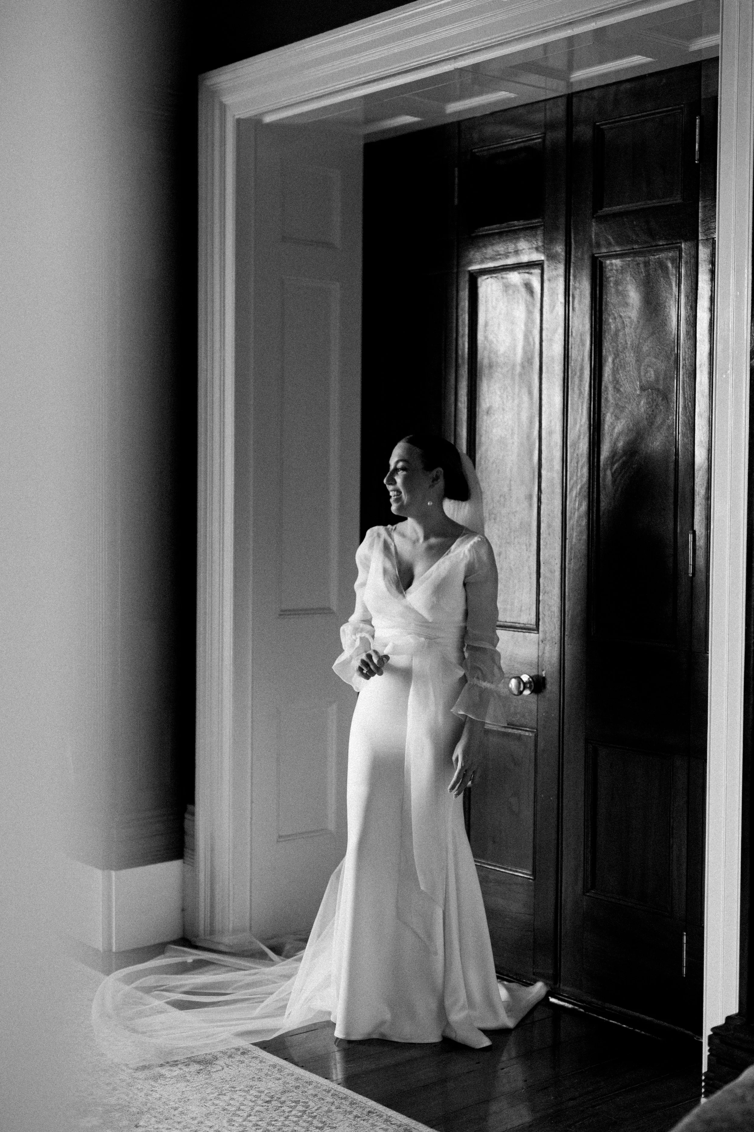 A bride in a wedding dress smiling and standing near a partially open wooden door in a room.