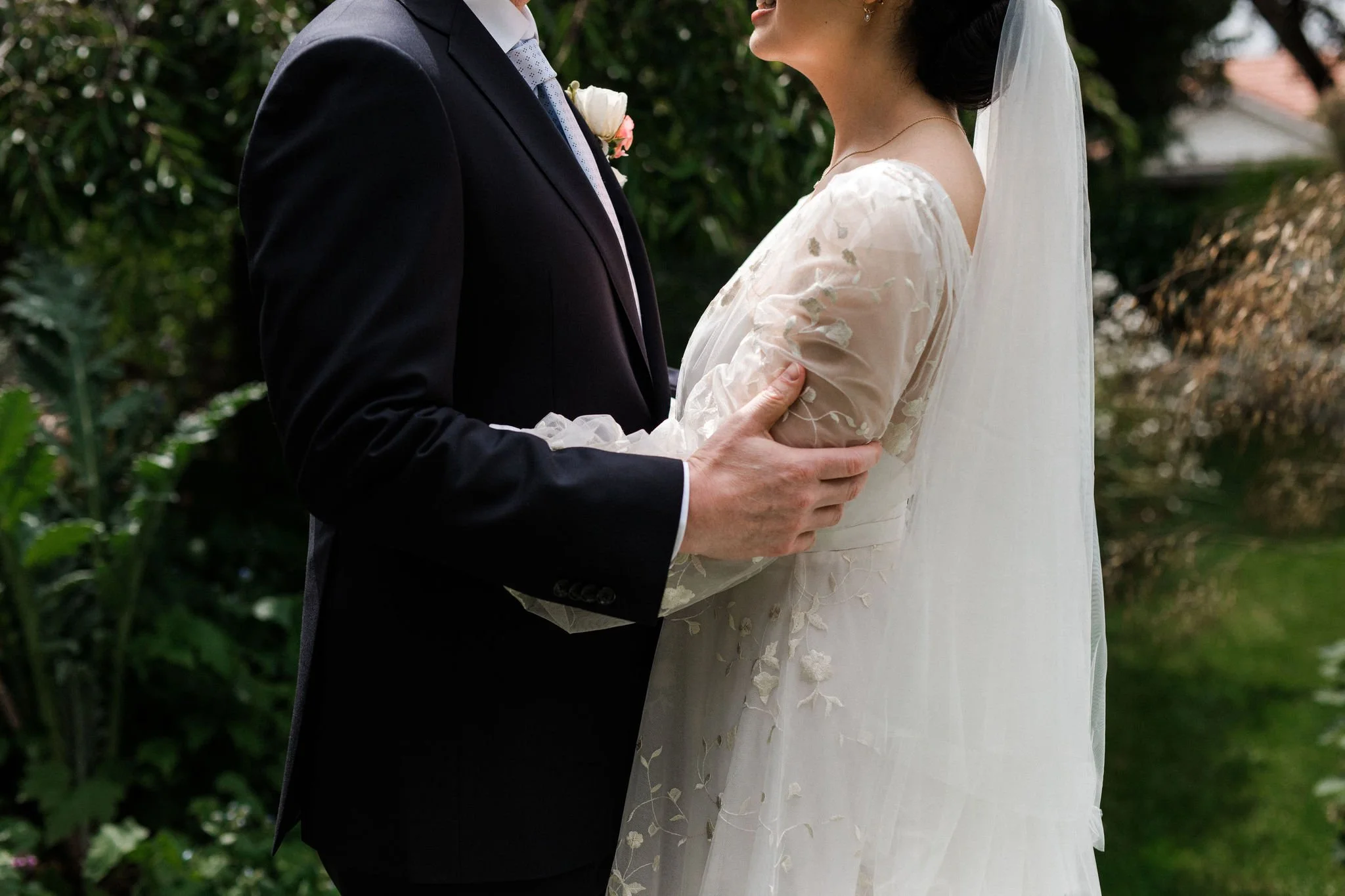 A bride and groom holding hands outdoors, with the groom wearing a black suit and the bride in a white wedding dress with floral lace details.