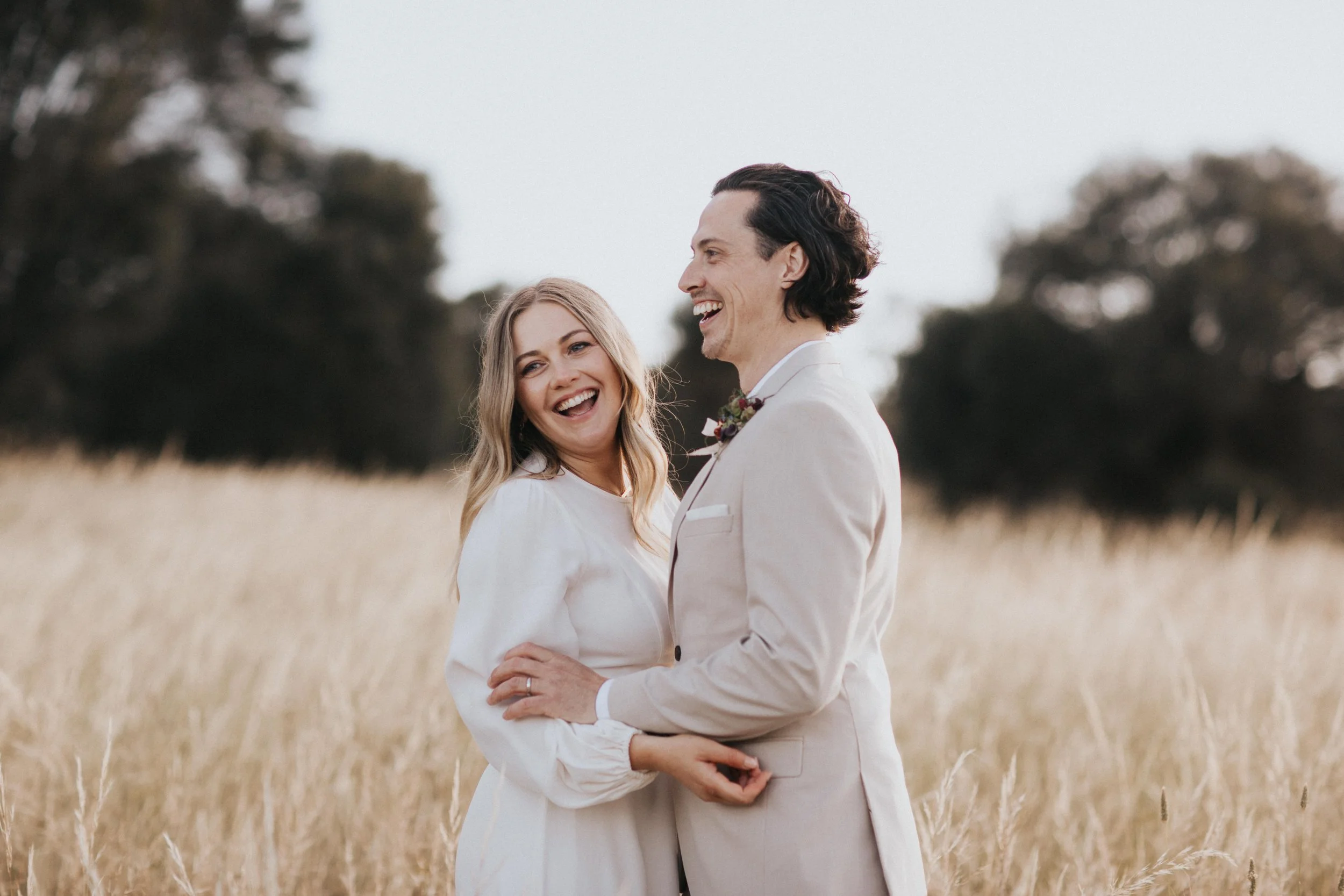 A smiling couple in wedding attire standing in a field of tall, dry grass, with trees in the background.