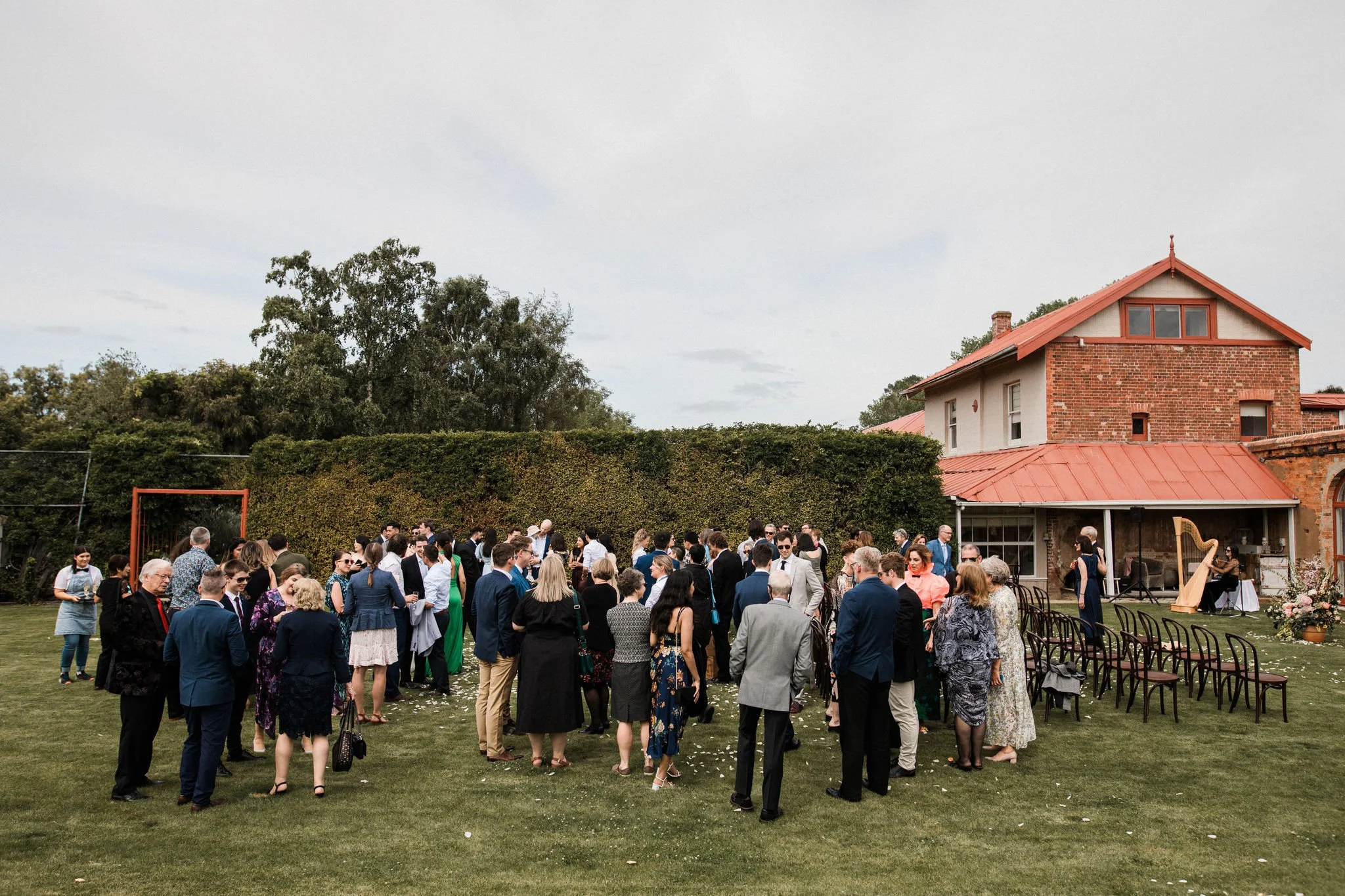 A large outdoor gathering of people dressed in formal attire on a lawn in front of a brick house with a harpist playing music.