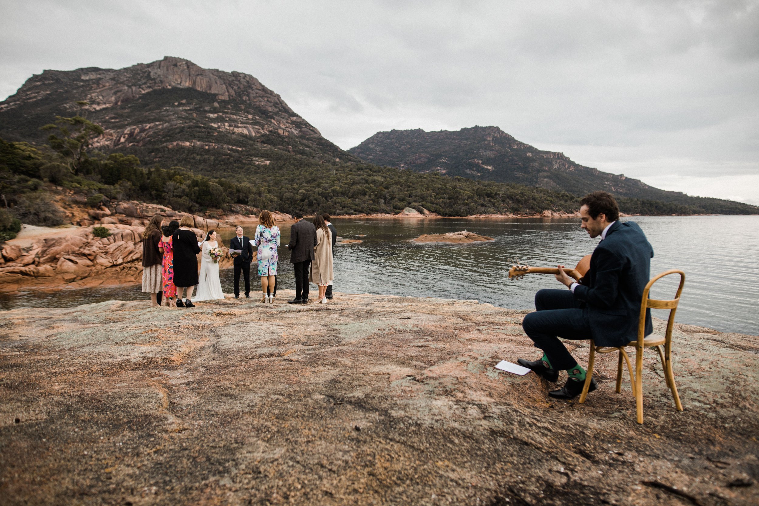 A wedding ceremony taking place on a rocky waterfront with mountains in the background, with a person playing guitar to the right.