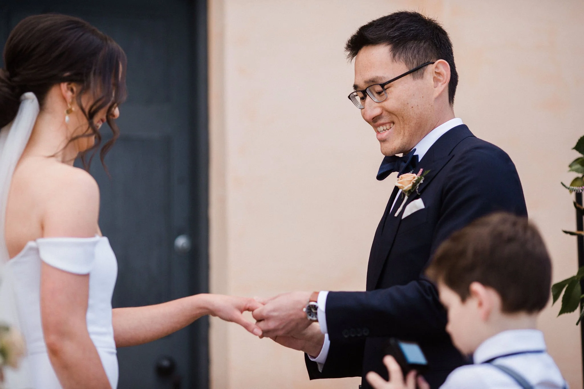 A groom in a dark suit and glasses placing a ring on a bride's finger during a wedding ceremony, with a young boy watching.