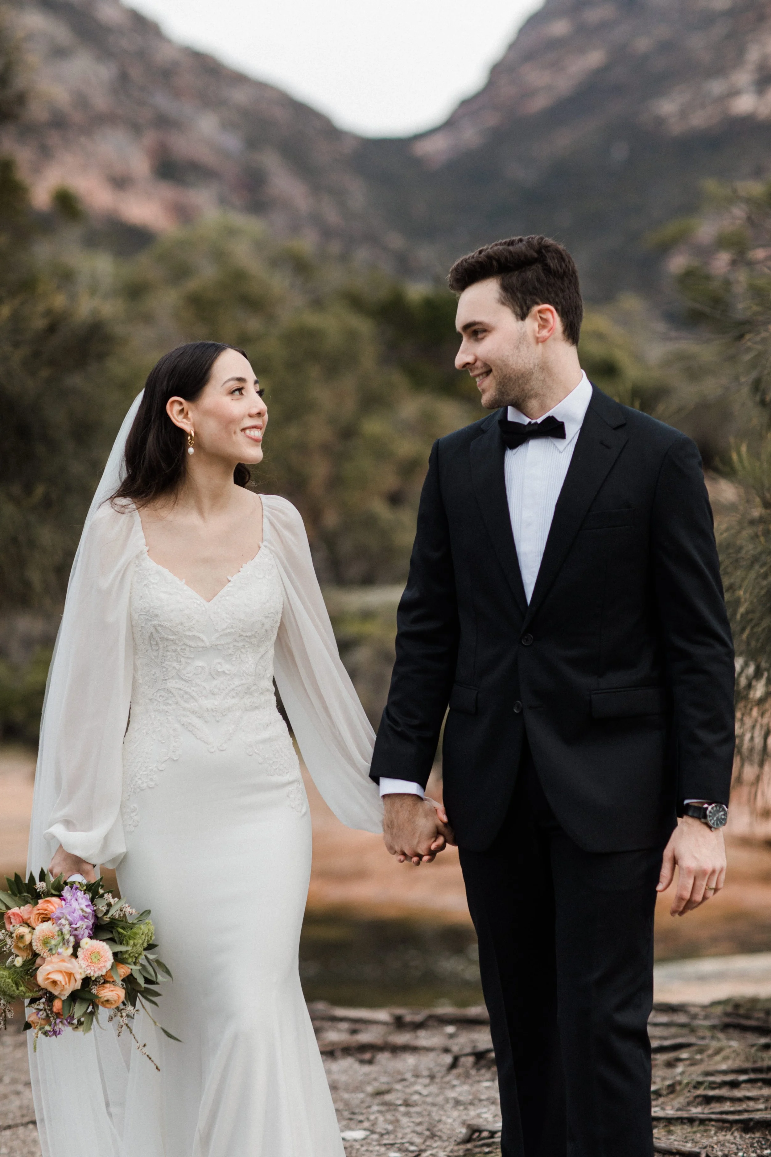 A bride and groom holding hands outdoors, smiling at each other, with mountains and trees in the background.