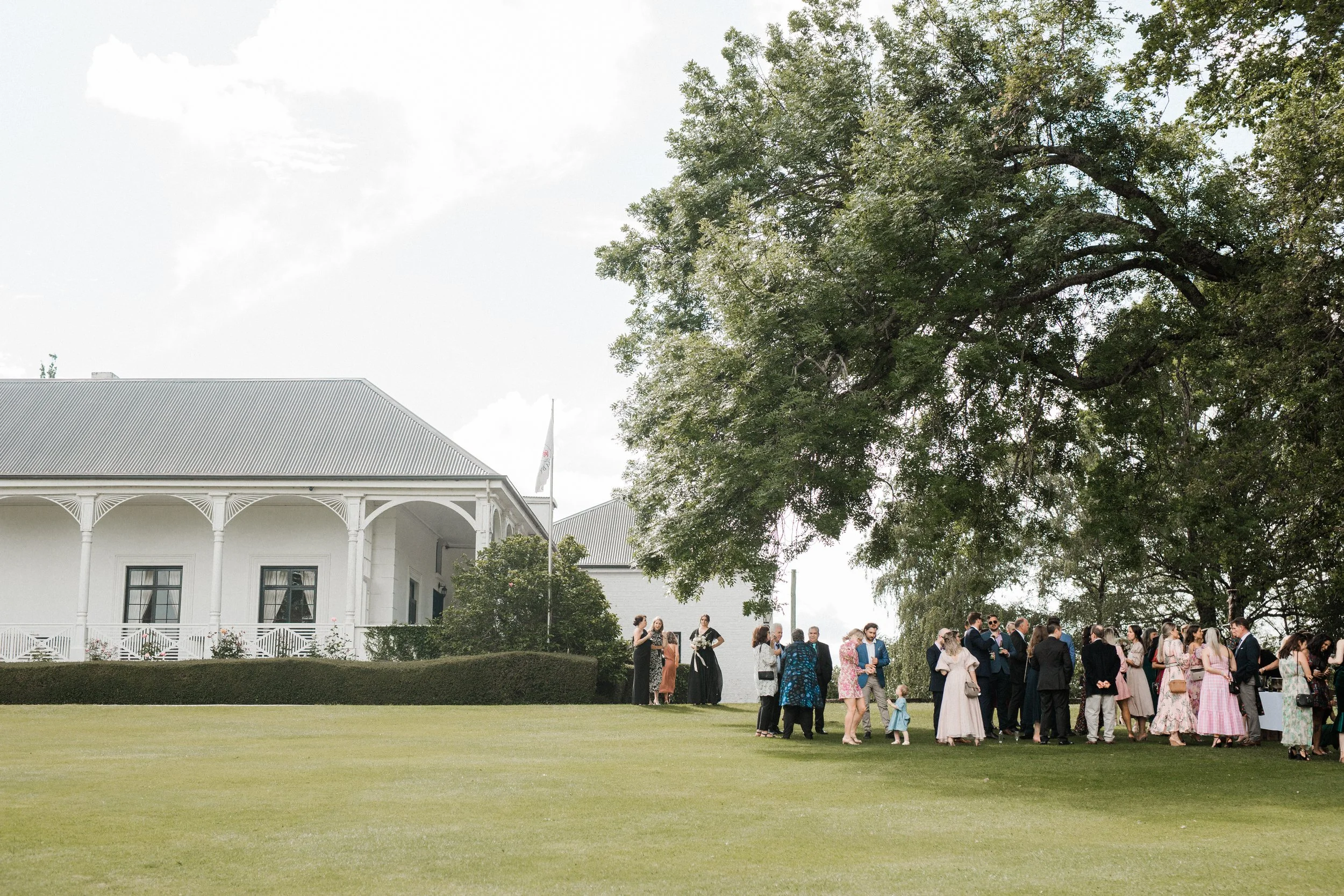 A large group of people gathered outdoors on a lawn near a white building with a veranda, tall trees in the background, and a partly cloudy sky.