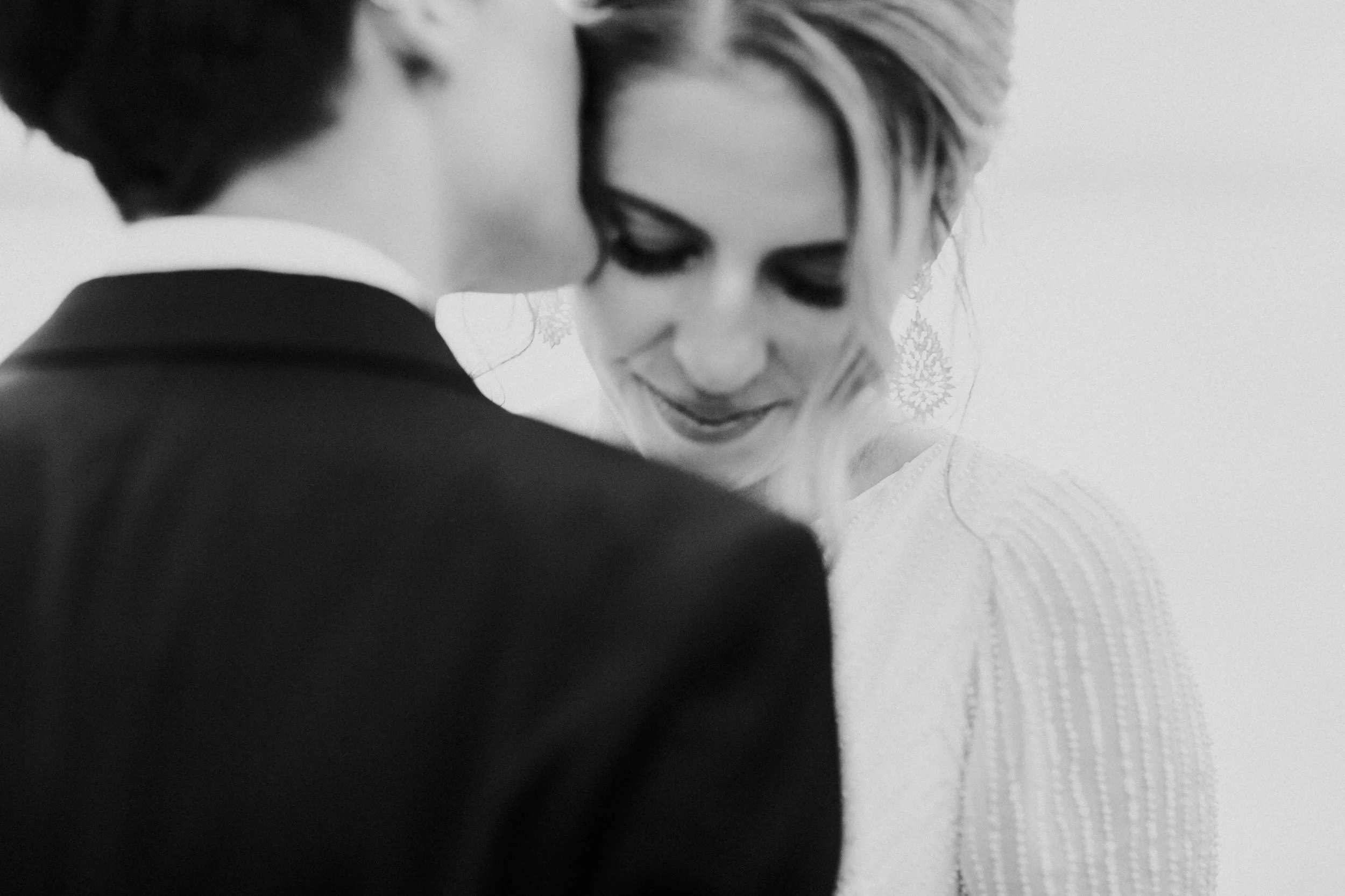 A black and white photo of two brides close together, touching foreheads and sharing an intimate moment, with one bride smiling softly.