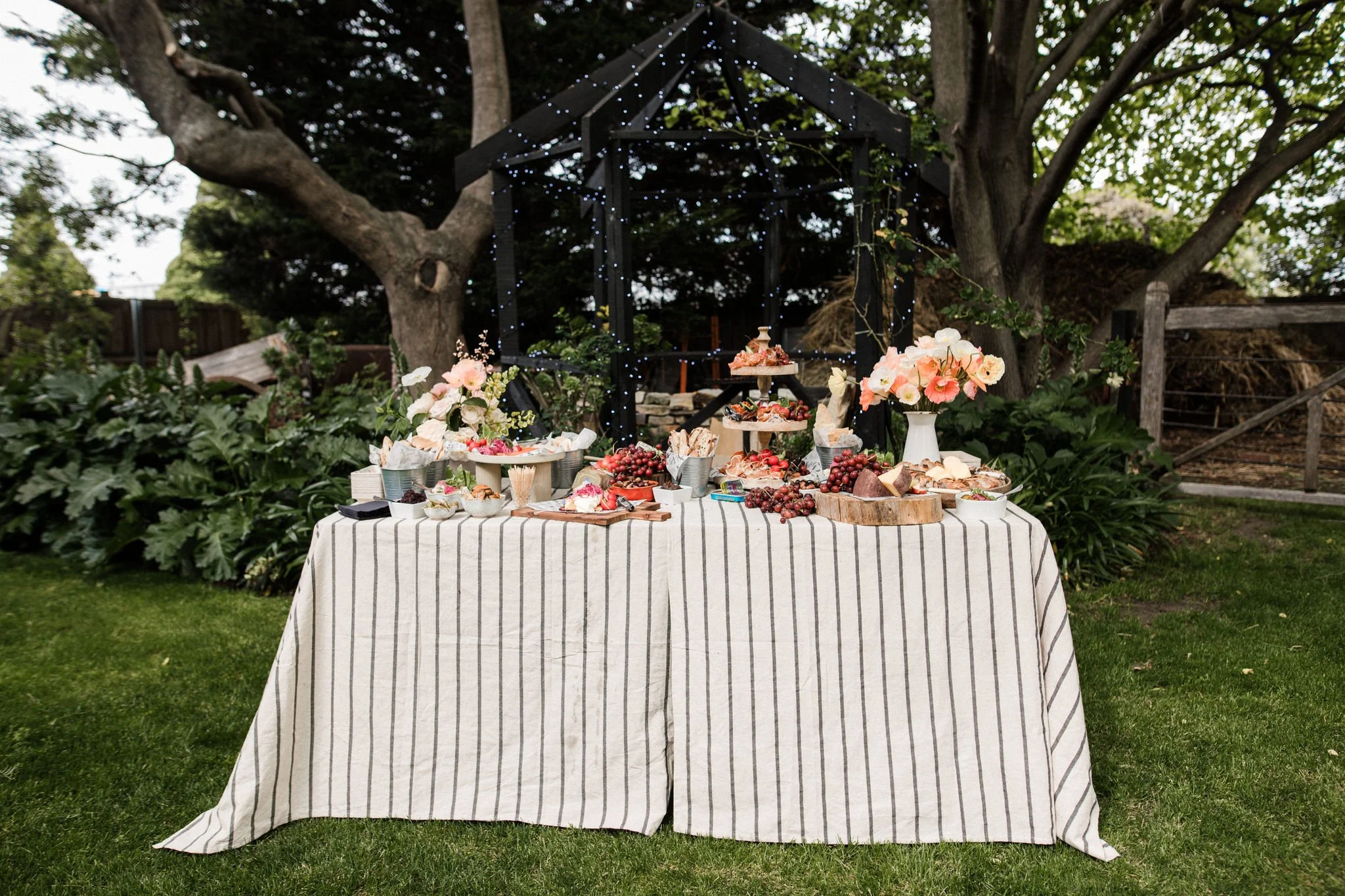 Outdoor dessert table with flowers, fruits, and cakes, set against trees with fairy lights.