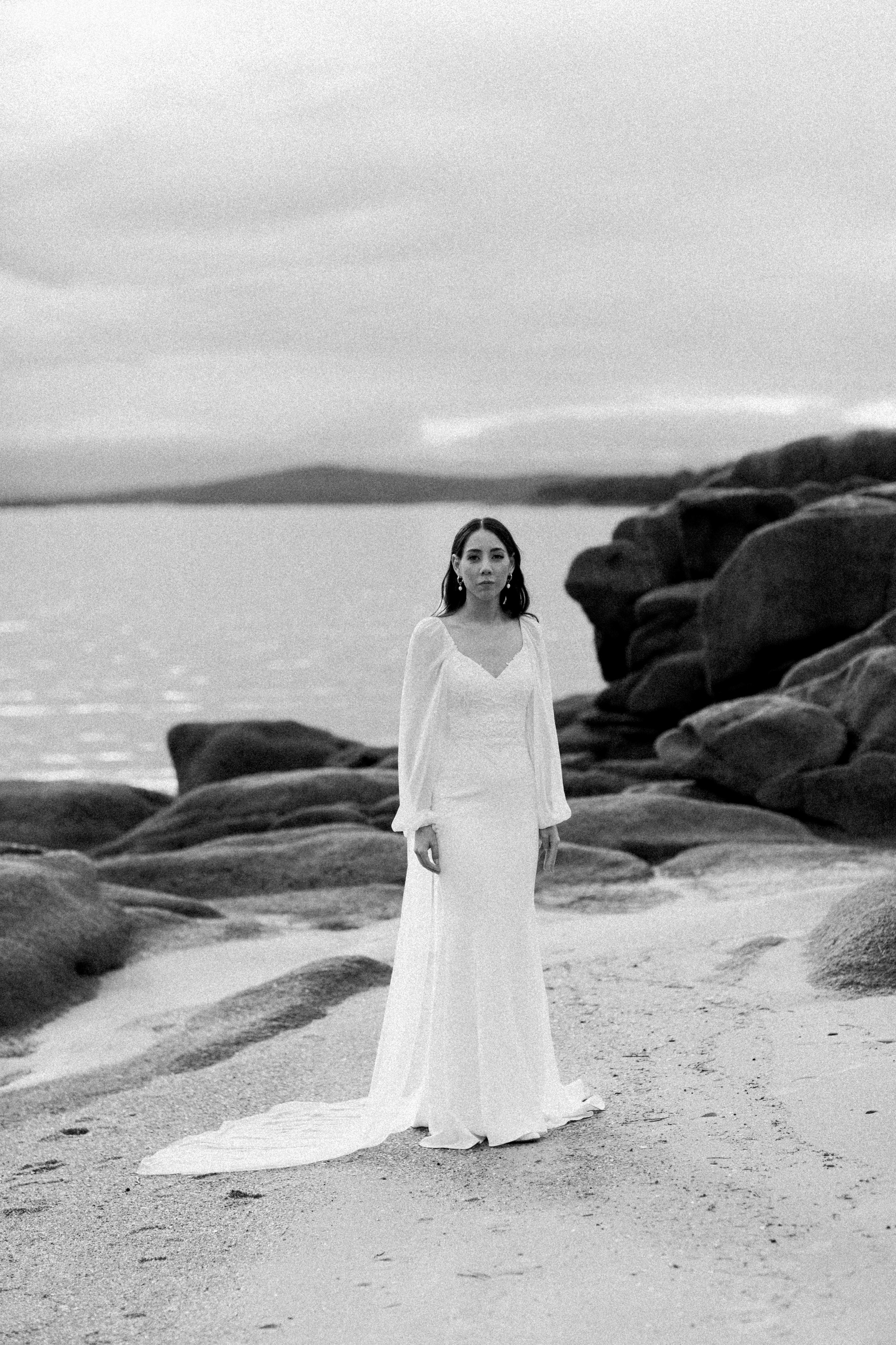 A woman in a white wedding dress standing on a sandy beach with rocks, water, and a cloudy sky in the background.