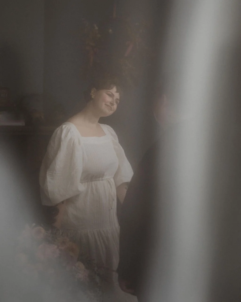 A woman smiling and looking through a frosted glass door, wearing a white dress with puffed sleeves.