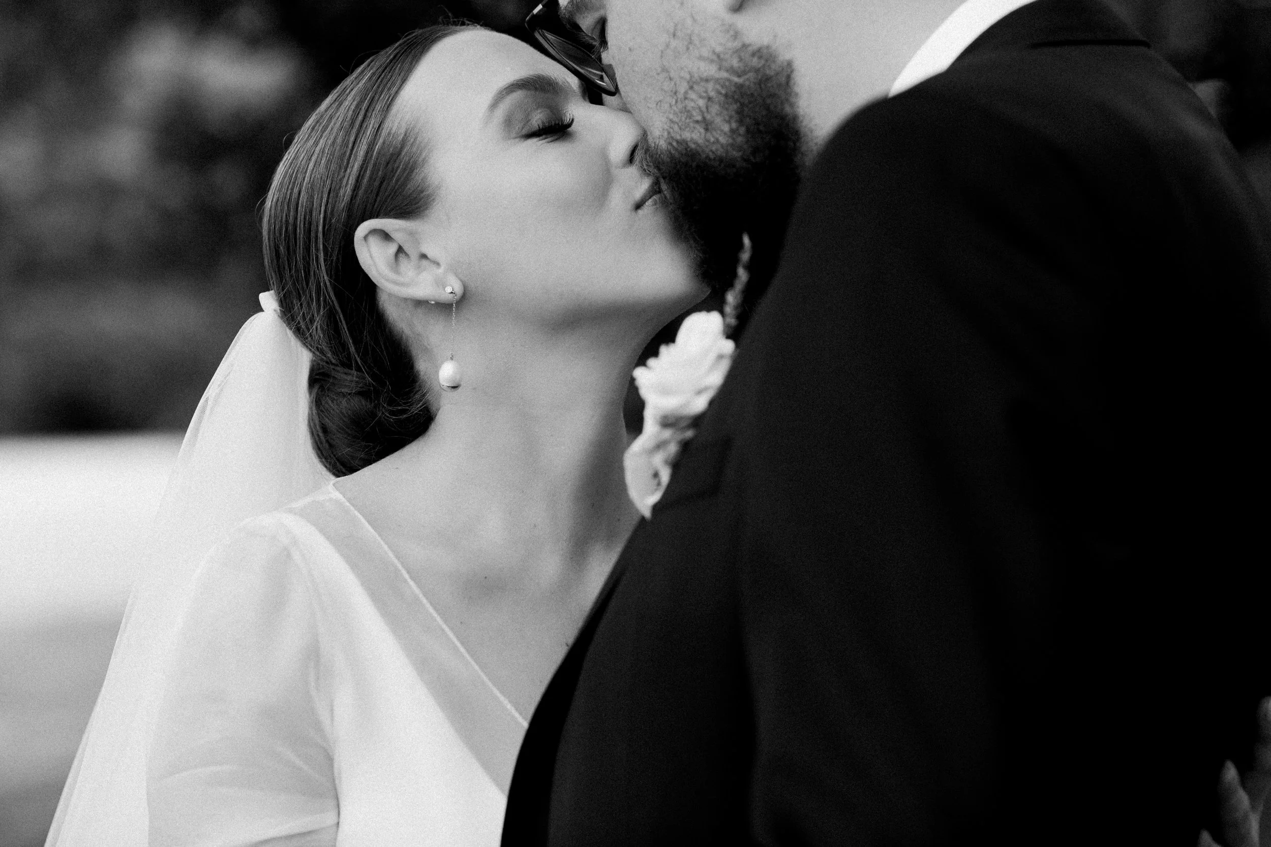 A black and white photo of a bride and groom sharing a kiss at their wedding, with the bride wearing earrings and a veil.