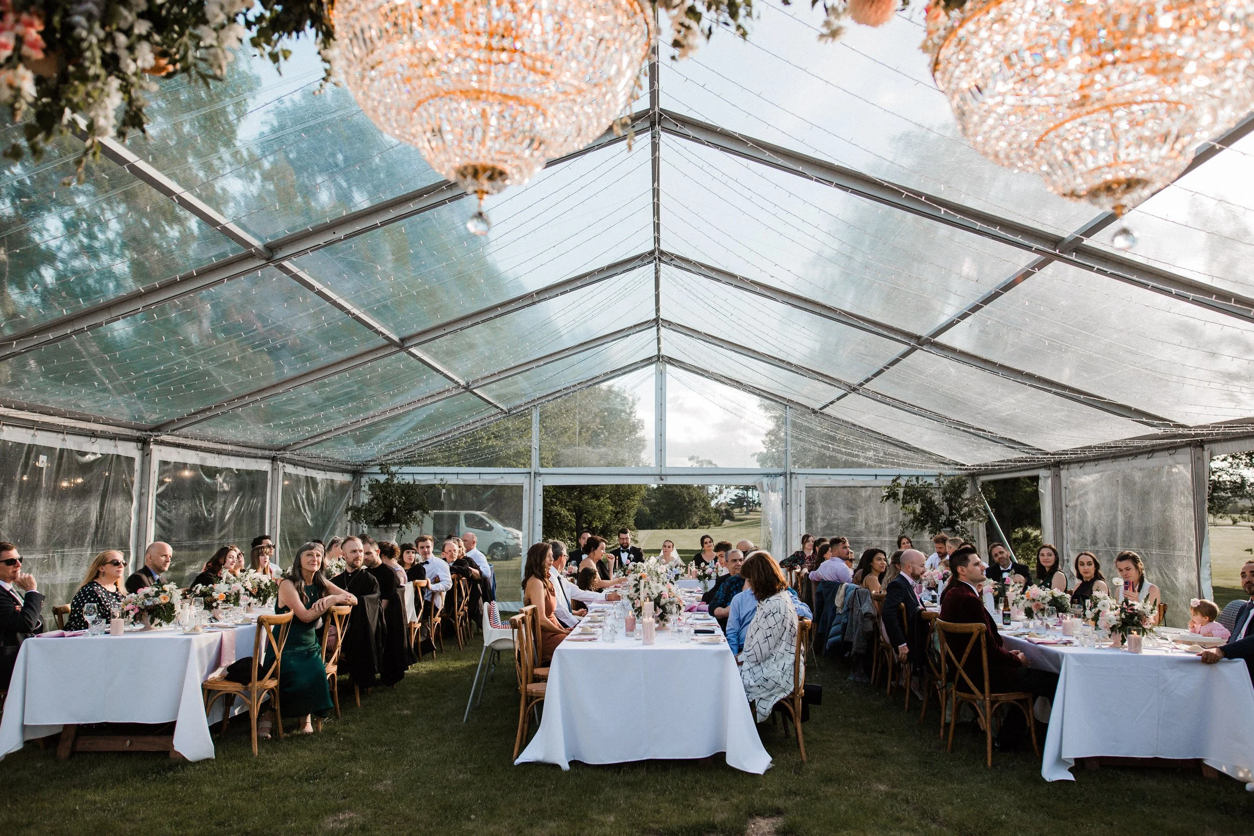 Guests seated at long wedding reception tables under a clear tent with chandeliers, floral centerpieces, and decorative lights outside.