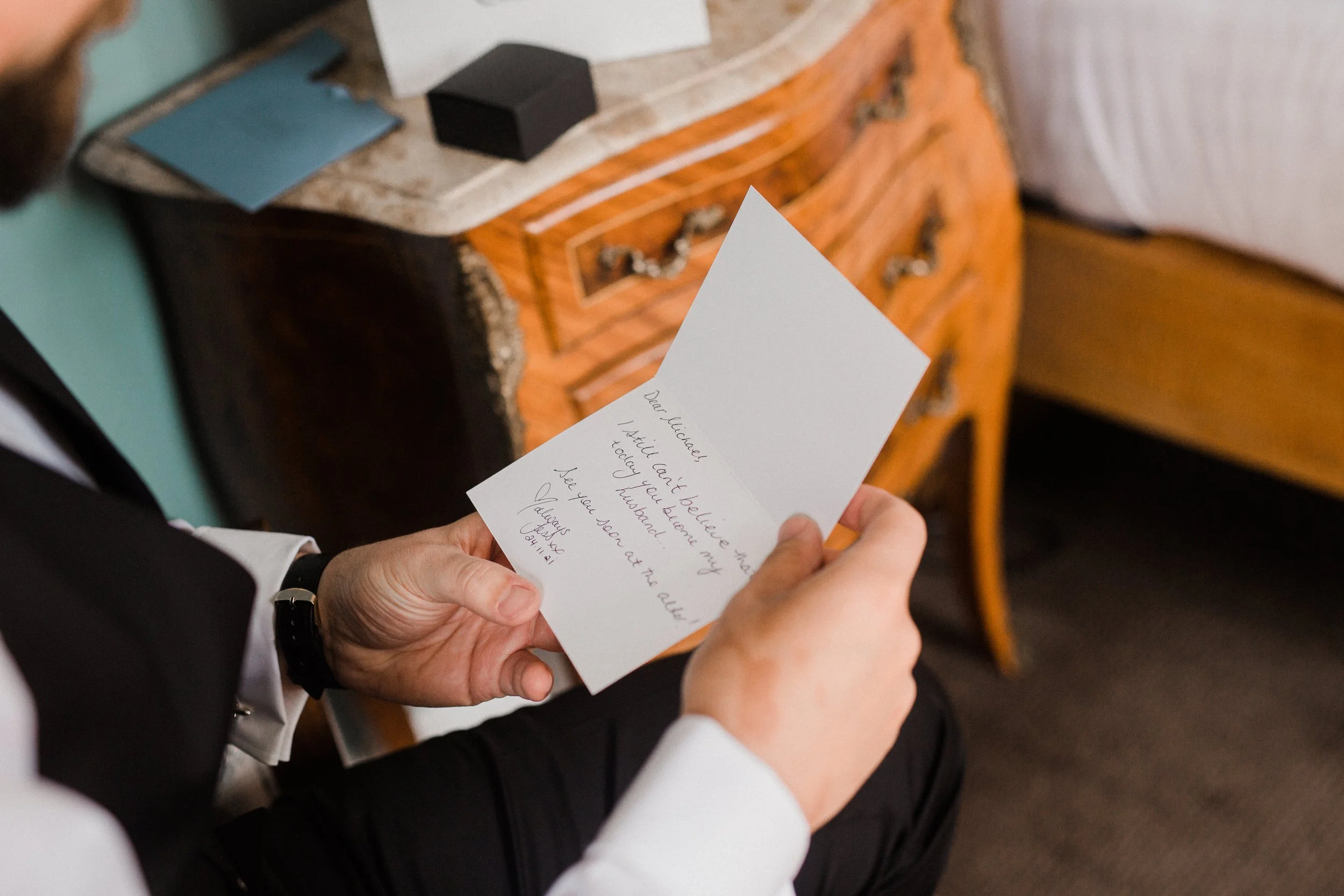 A person in a suit holding a handwritten note, sitting next to a wooden dresser and a bed in a cozy room.
