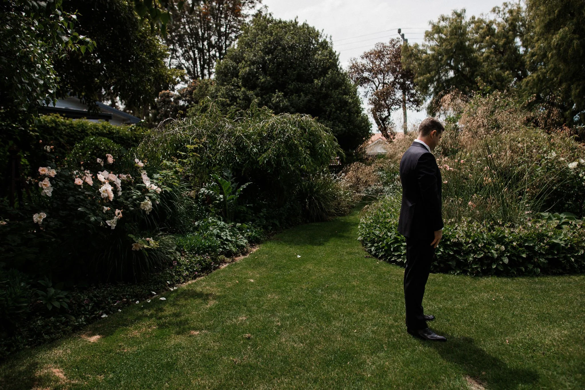 A man in a black suit stands alone on a lush green lawn in a garden filled with flowering bushes and trees.