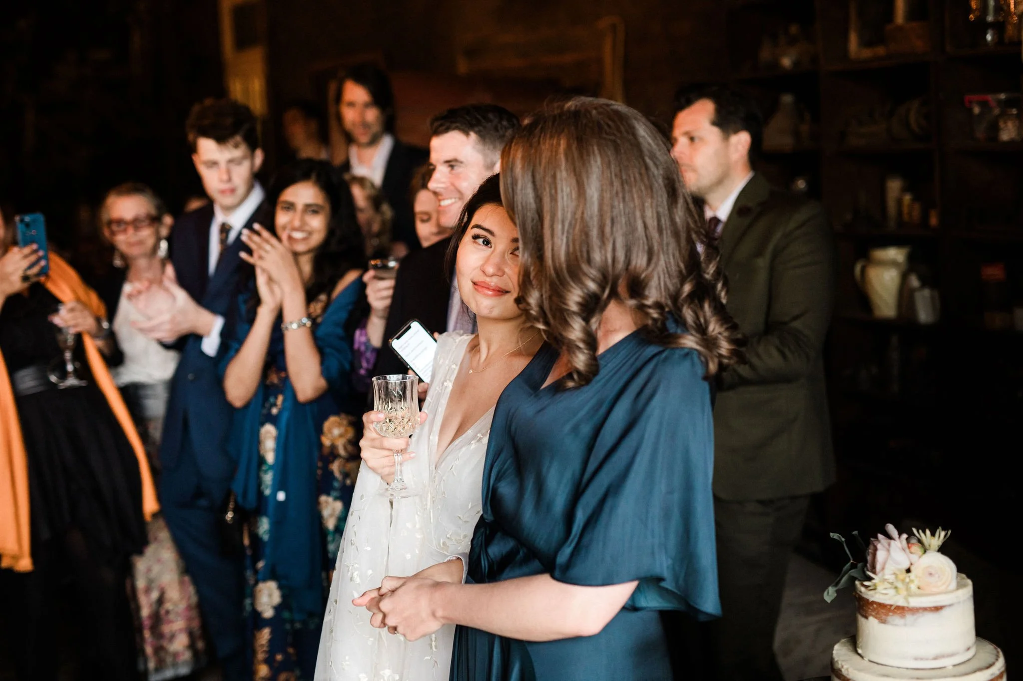 A woman in a white dress holding a glass of wine is looking at another woman in a blue dress, surrounded by a group of people at a celebration or wedding reception.