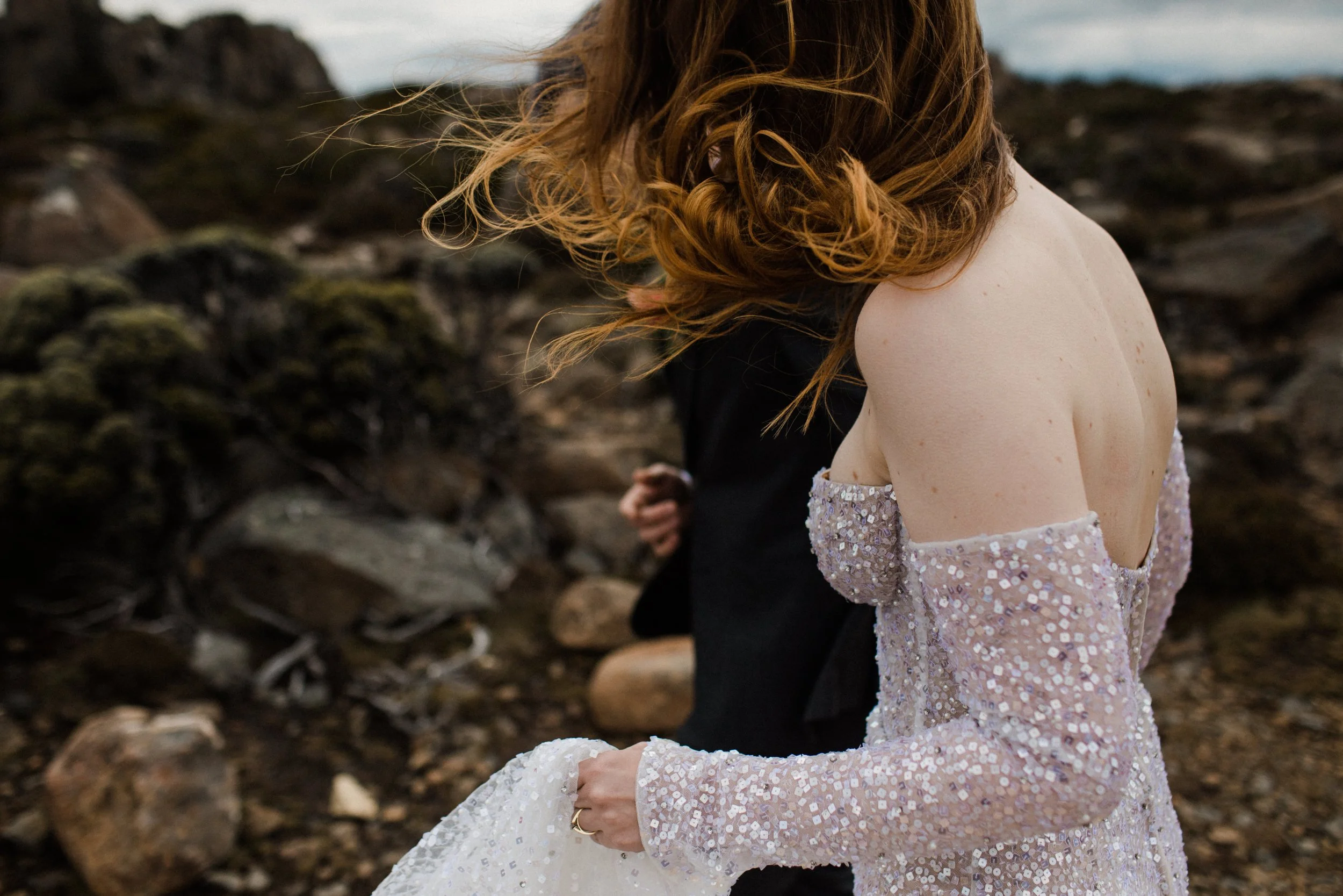 A woman with curly red hair wearing an off-shoulder sequined dress stands near rocks outdoors.