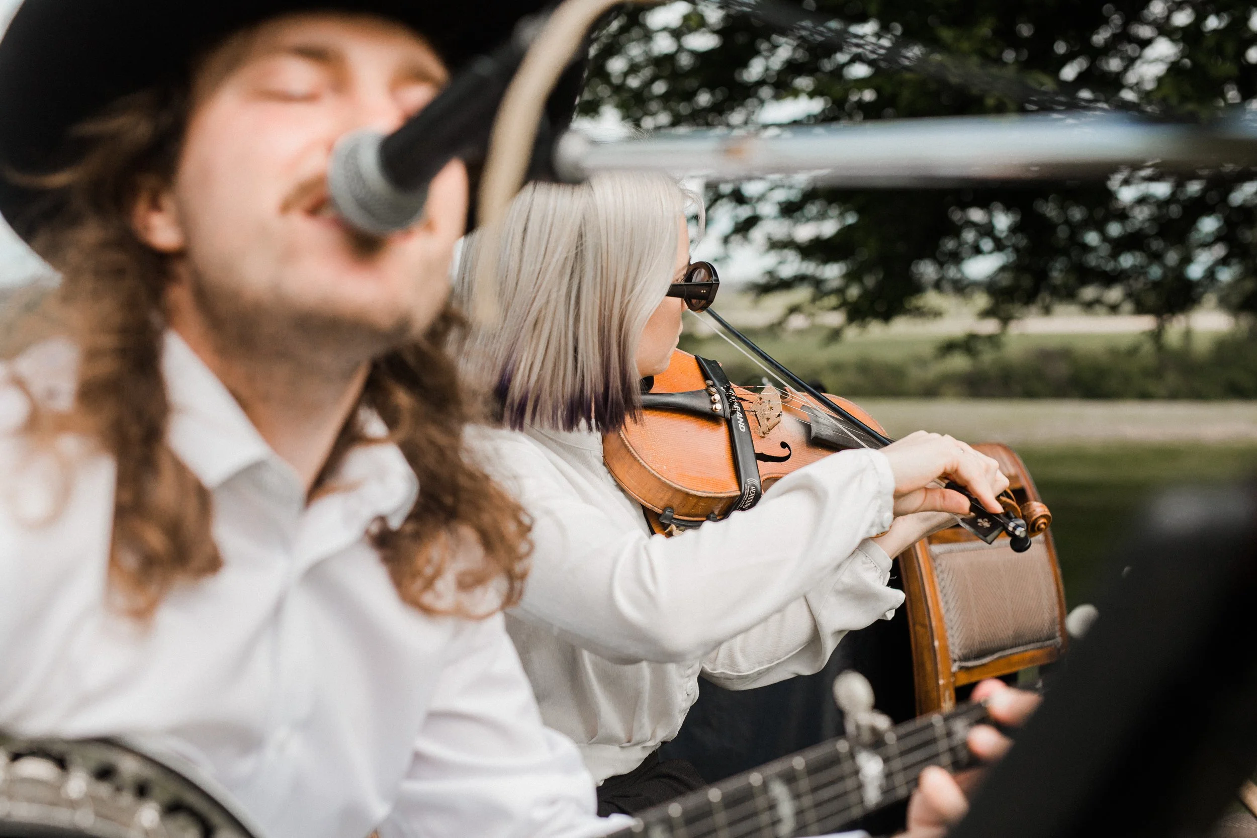Two musicians outdoors: one playing a guitar with curly hair and the other playing a violin with blonde hair and sunglasses, blurred woman in the foreground.