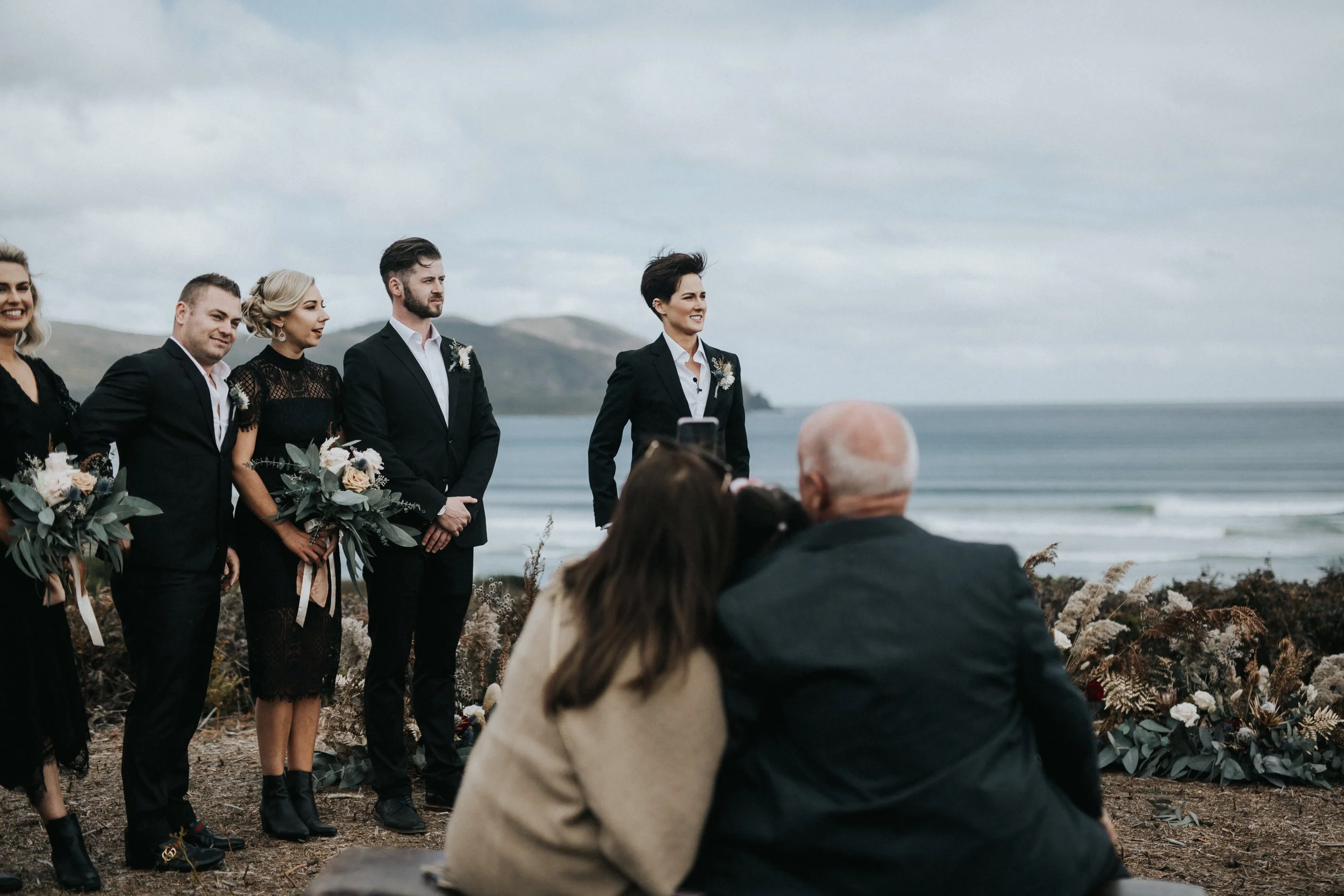 A wedding ceremony taking place on a beach with the ocean and hills in the background, featuring a group of people dressed in formal black attire.