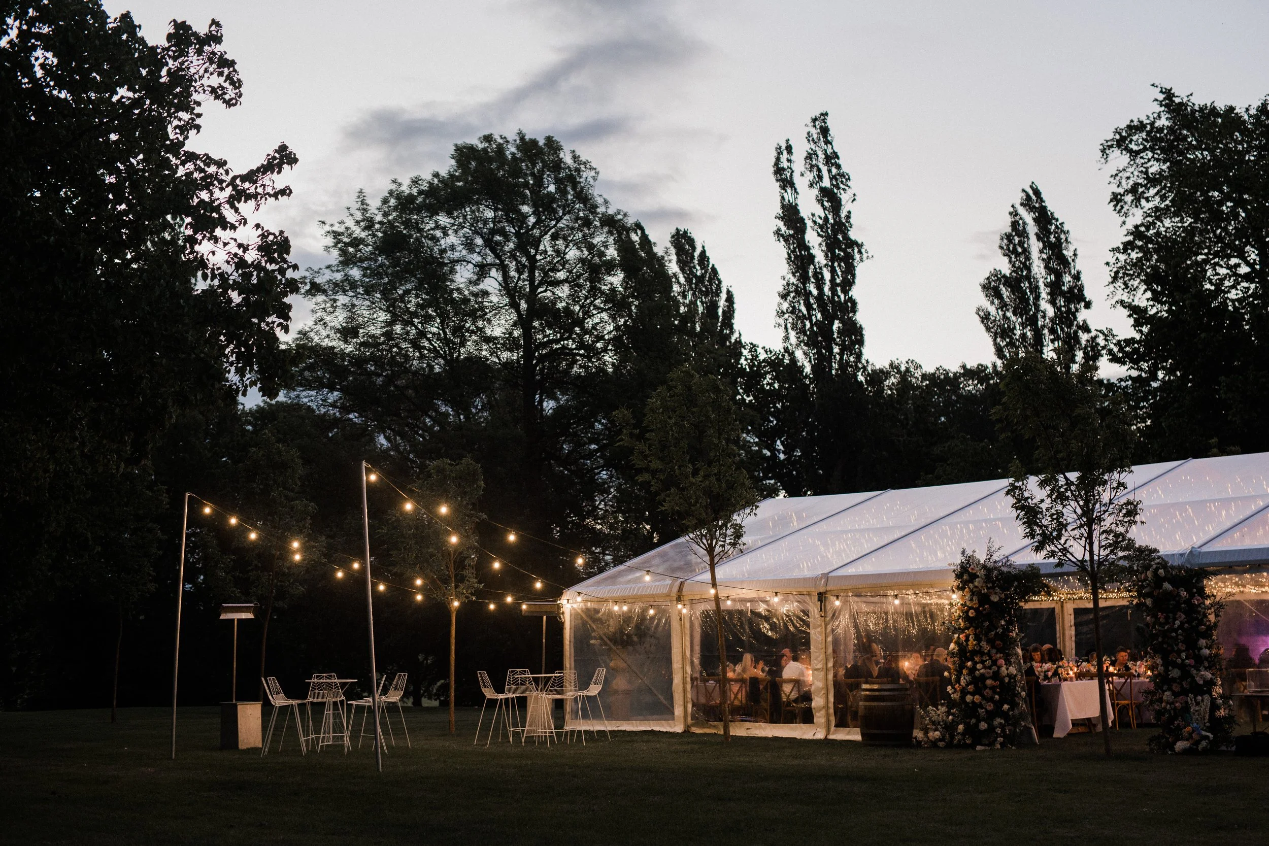 An outdoor evening event with a large white tent decorated with string lights, surrounded by trees and open grassy area, with tables and chairs inside and outside.