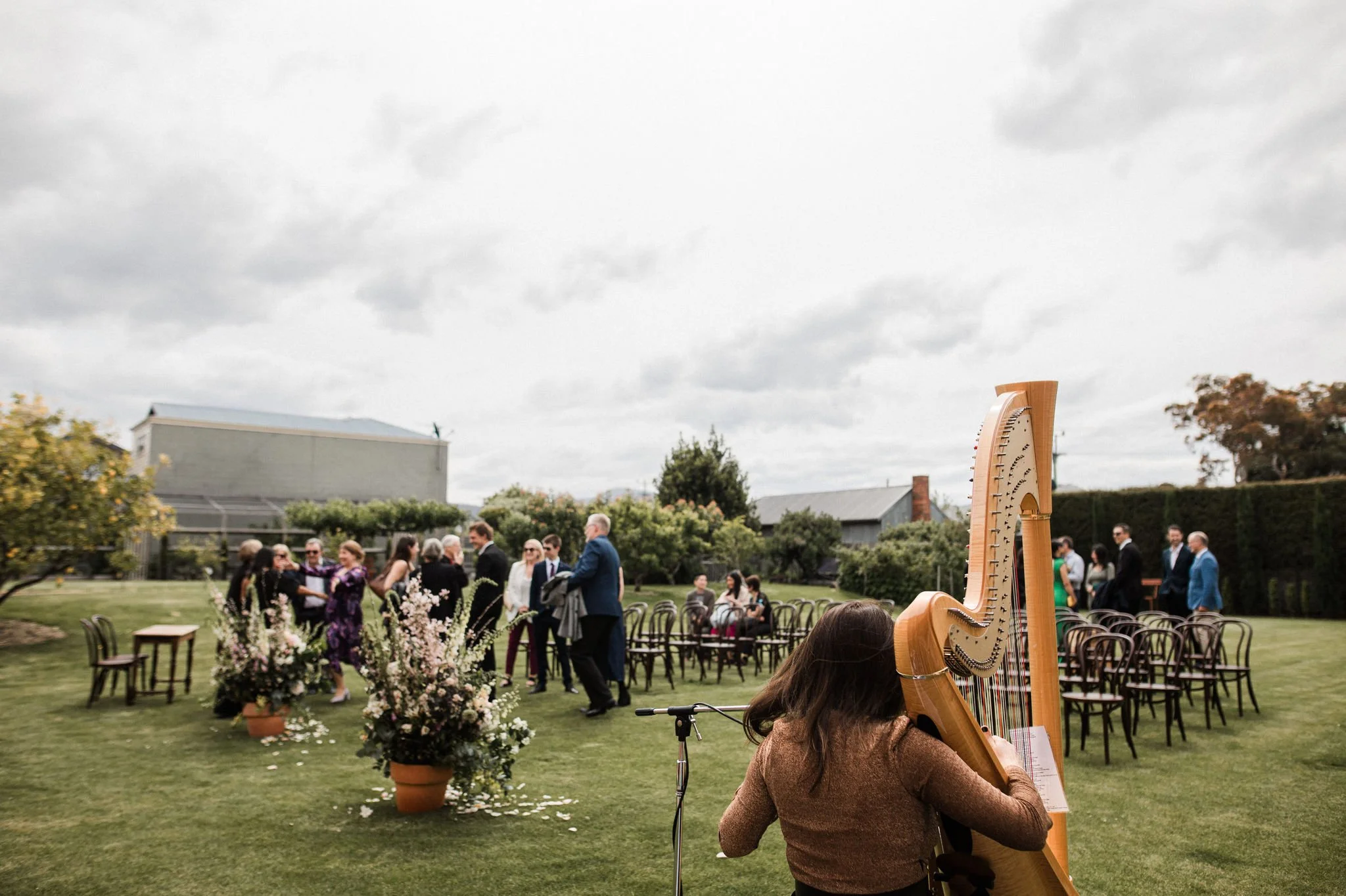 Outdoor wedding ceremony with people mingling, seated guests, and a harpist playing on a grassy lawn, decorated with flower arrangements and chairs arranged for the event.