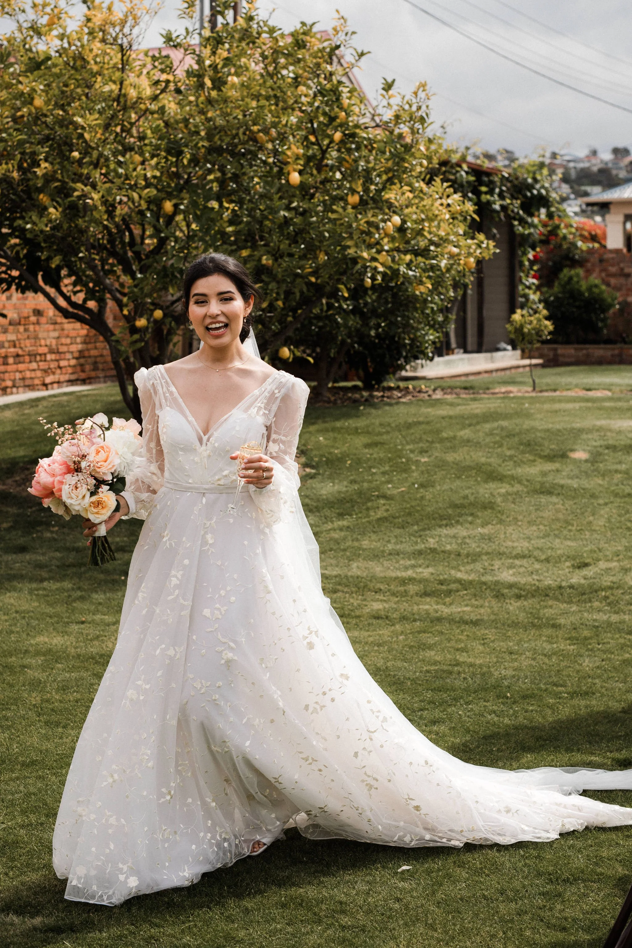 A smiling bride in a white wedding dress holding a bouquet of pink and white flowers, standing on a grassy lawn with a lemon tree and houses in the background.