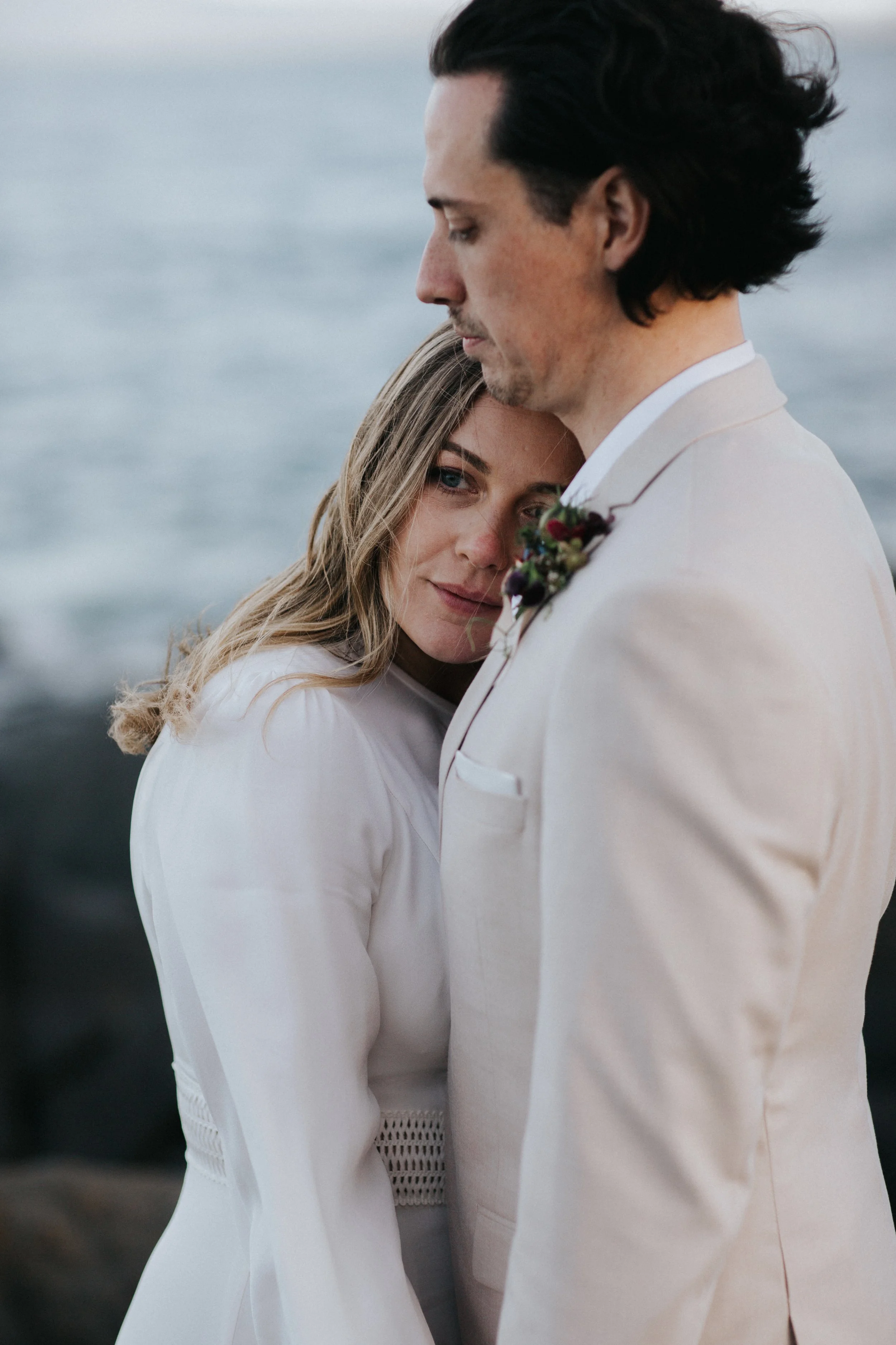 A woman with long blonde hair and blue eyes leaning her head on a man's chest, who has black hair. They are dressed in formal wedding attire, standing outdoors near the water.