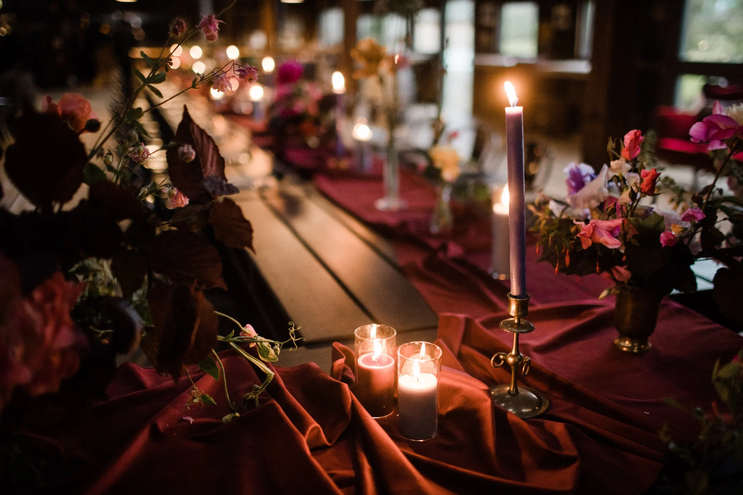Elegant dining table with lit candles, surrounded by pink and purple flowers, set with burgundy tablecloths in a dimly lit, cozy room.