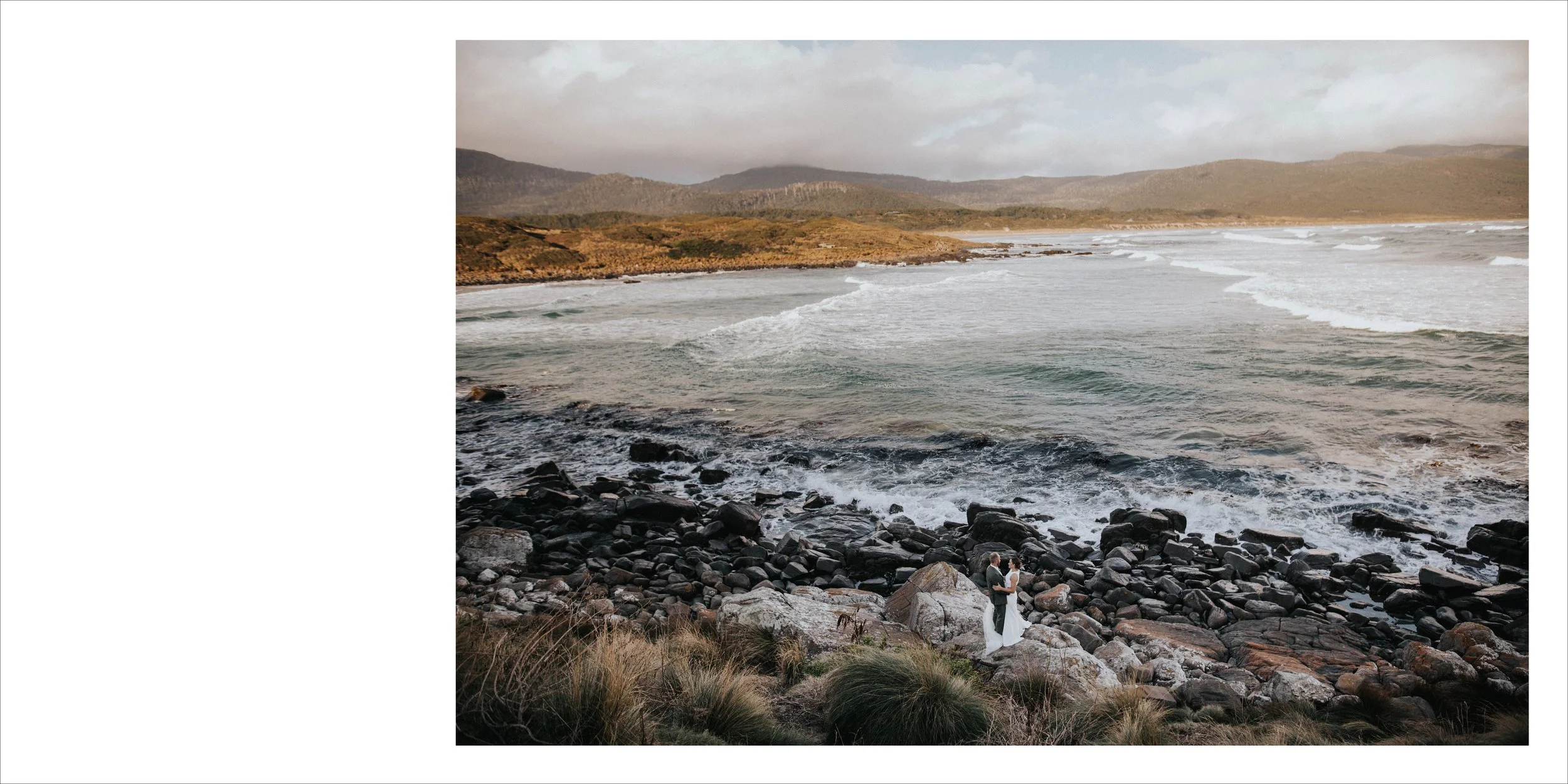 A bride and groom standing on a rocky shoreline by the ocean, with waves crashing and a landscape of hills in the background.