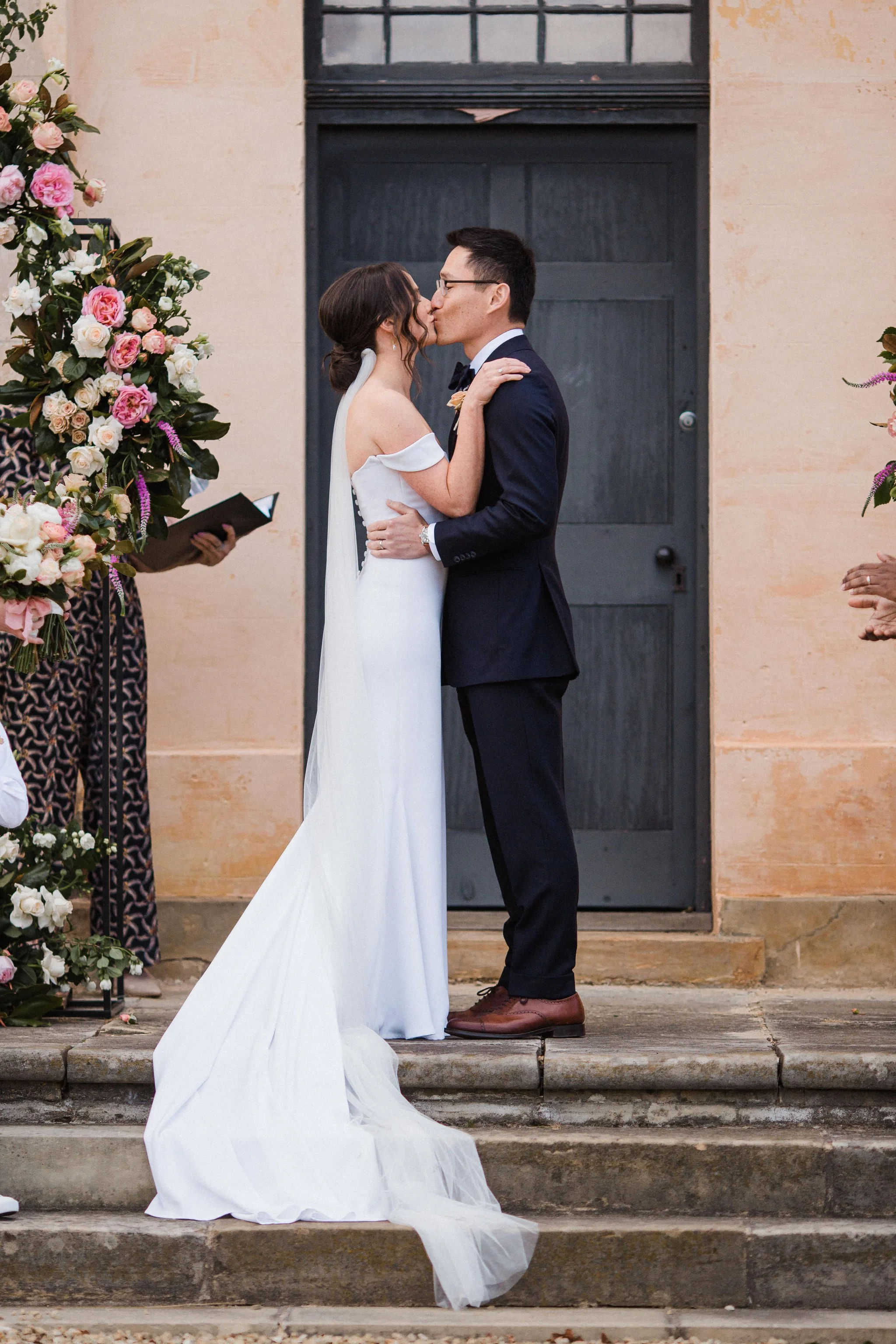 A bride and groom kiss during a wedding ceremony on stone steps outside a building, with floral arrangements on the side.